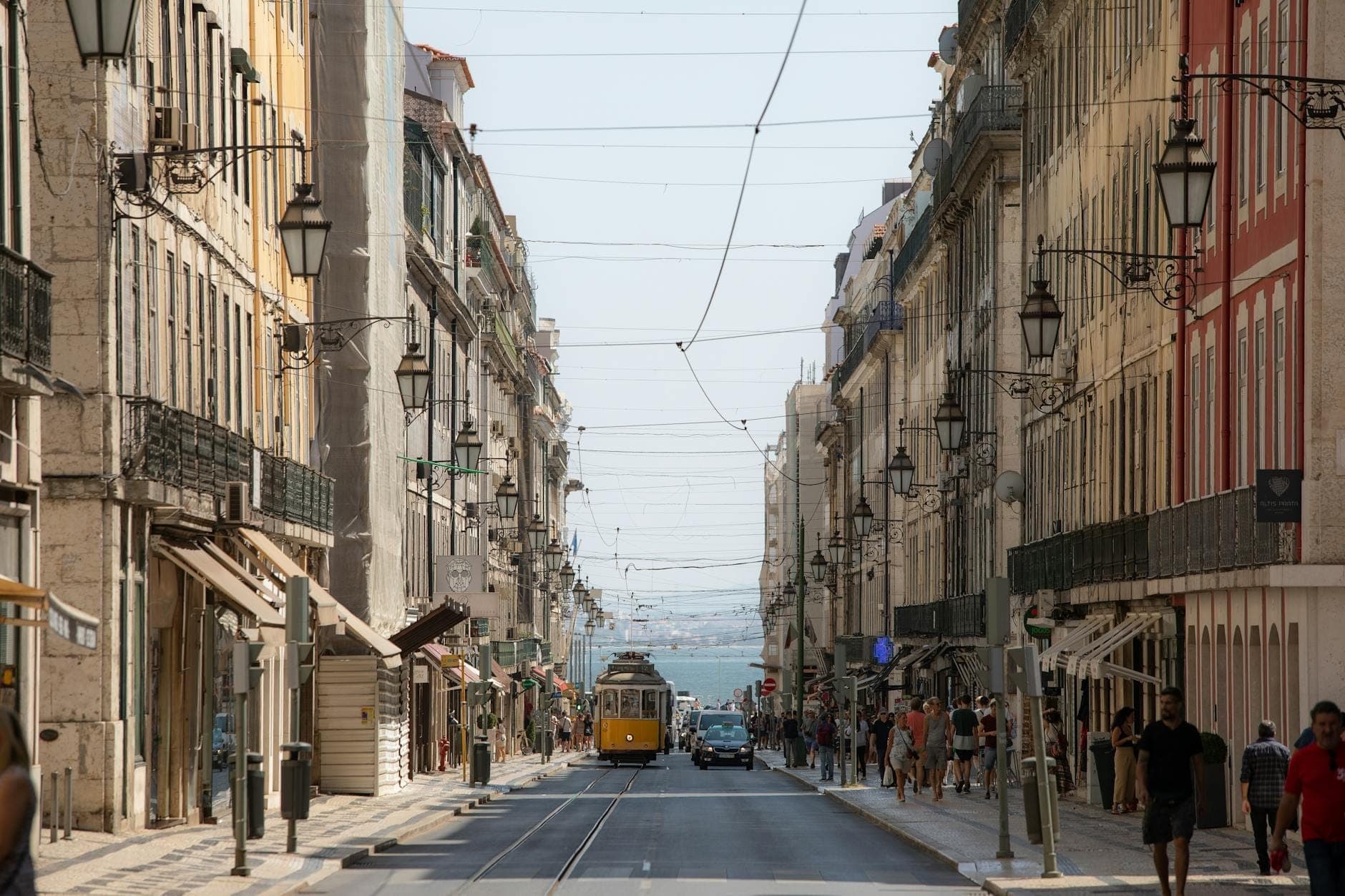 Ampla rua de Lisboa com eléctrico clássico, edifícios históricos e luzes festivas suspensas num dia ensolarado.
