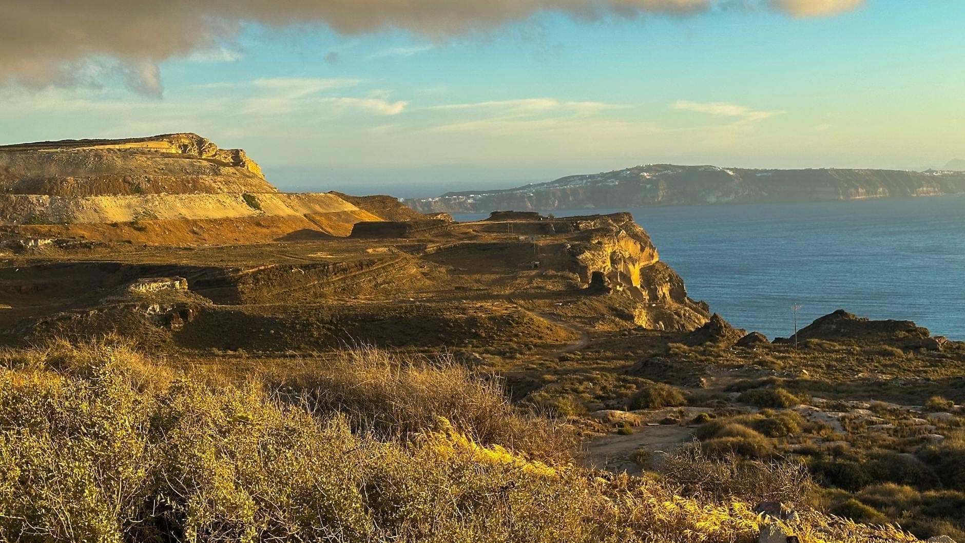 Paesaggio costiero di Santorini con scogliere frastagliate, terreno giallo-marrone, vegetazione rada e Mar Egeo, che evoca percorsi verso Akrotiri e Mesa Vouno.