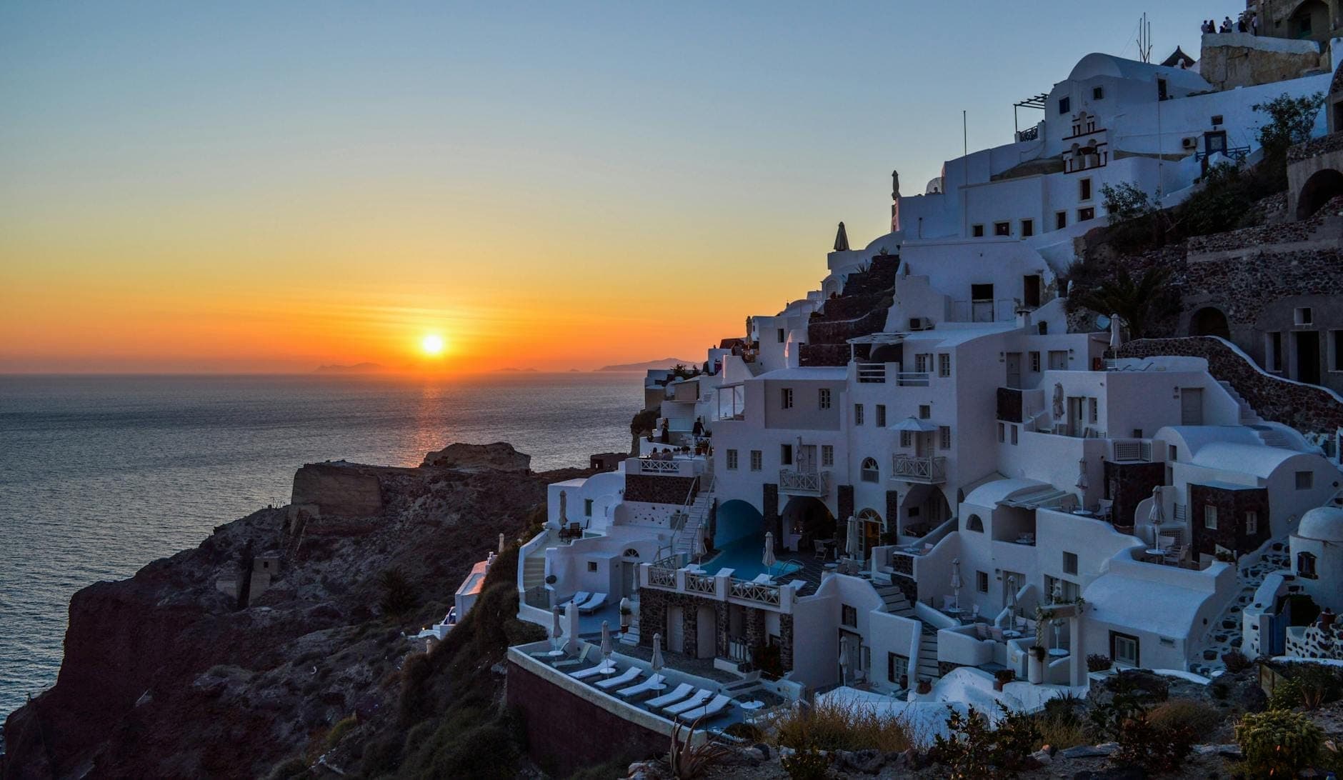 Edifici sul ciglio della scogliera a Santorini con vista sul mare al tramonto, con il sole basso all'orizzonte, che cattura una classica vista del tramonto.