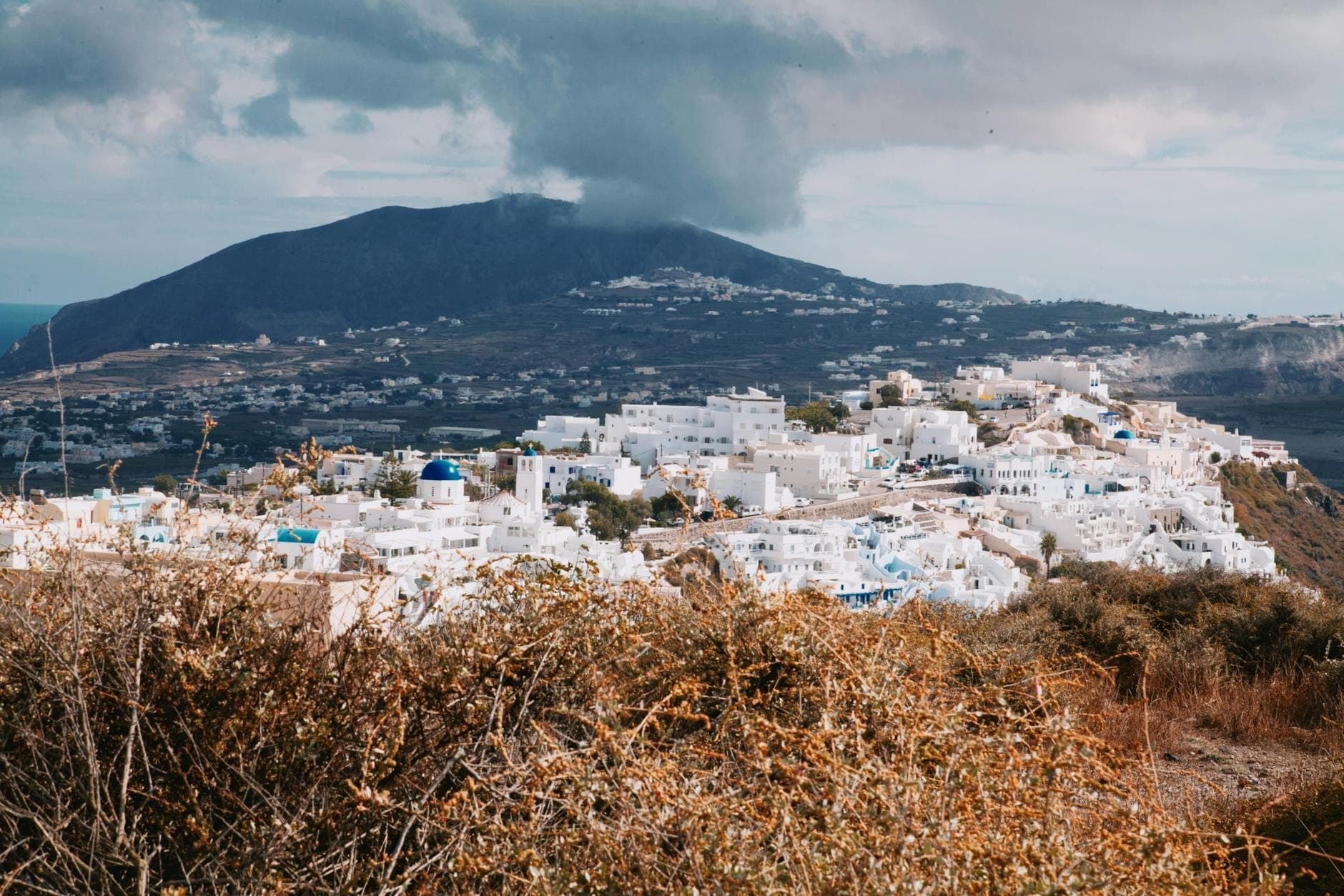 Villaggio in cima a una collina a Santorini con edifici bianchi e sfondo montuoso, circondato da paesaggio rurale e insediamenti tradizionali.