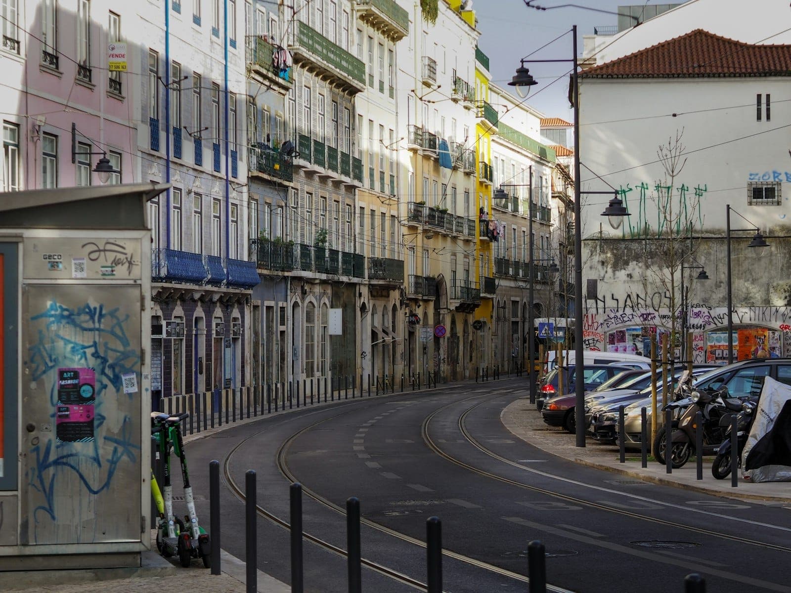 Rua curvilínea com trilhos de bonde em Santos ou Cais do Sodré, em Lisboa, com prédios históricos coloridos, arte de rua, scooters estacionadas e luz dourada do fim de tarde.