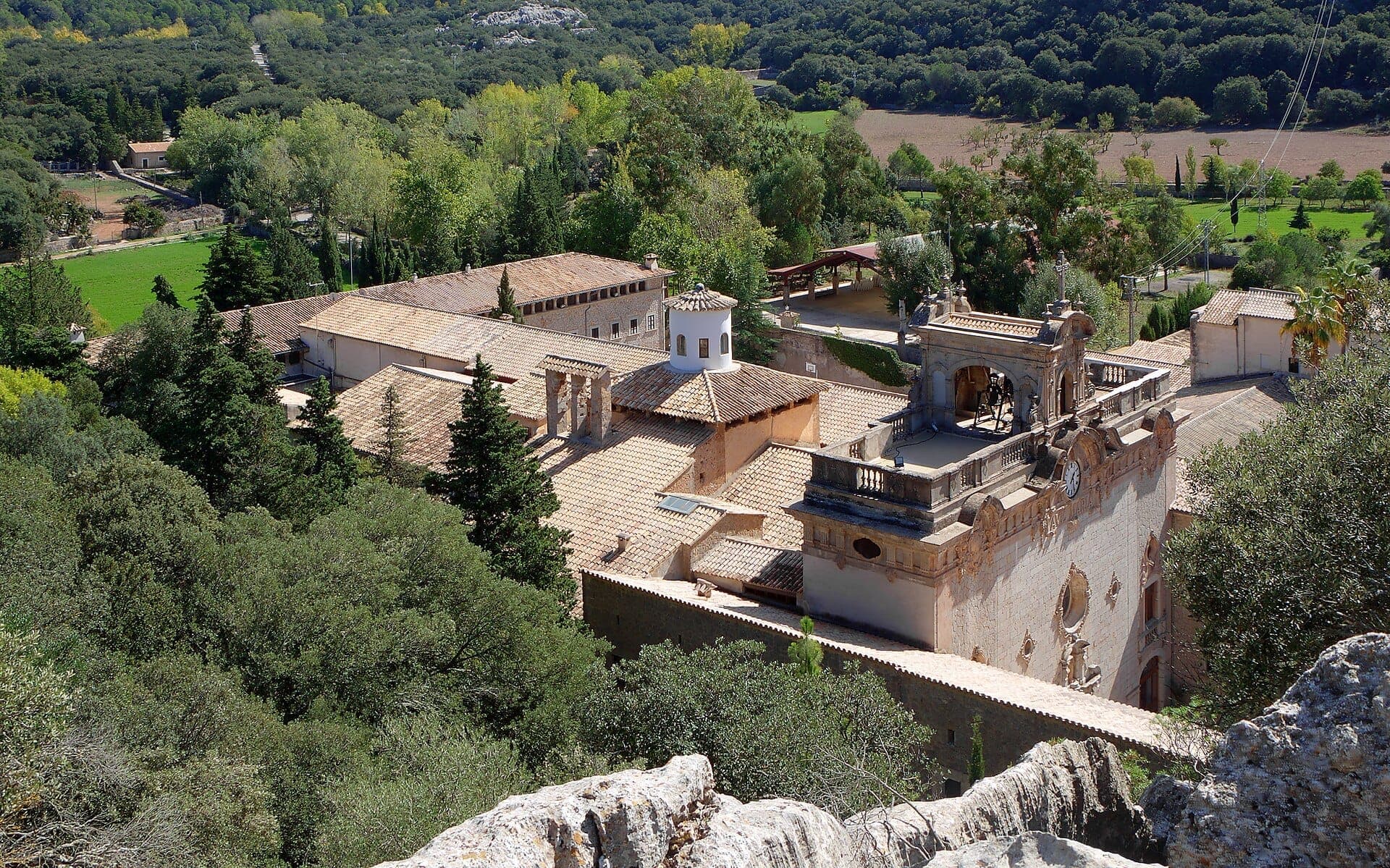 Luftaufnahme des Santuari de Lluc mit Terrakottadächern, Uhrturm und üppigem grünen Wald in den Tramuntana-Bergen.