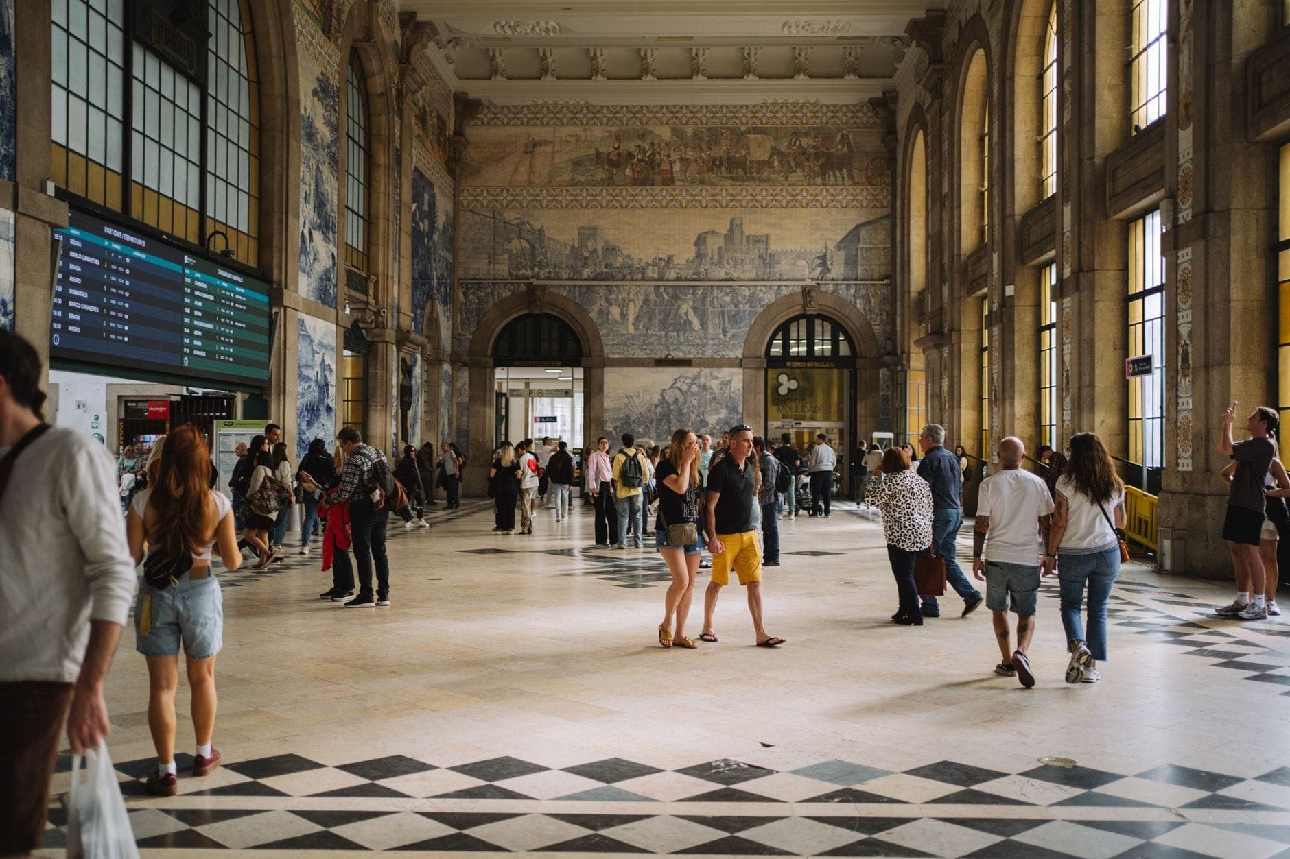 Innenansicht des Bahnhofs São Bento mit großen Bogenfenstern, kunstvollen Azulejo-Wandgemälden, einem Schachbrettboden und Besuchern, die die historischen Kunstwerke bestaunen.