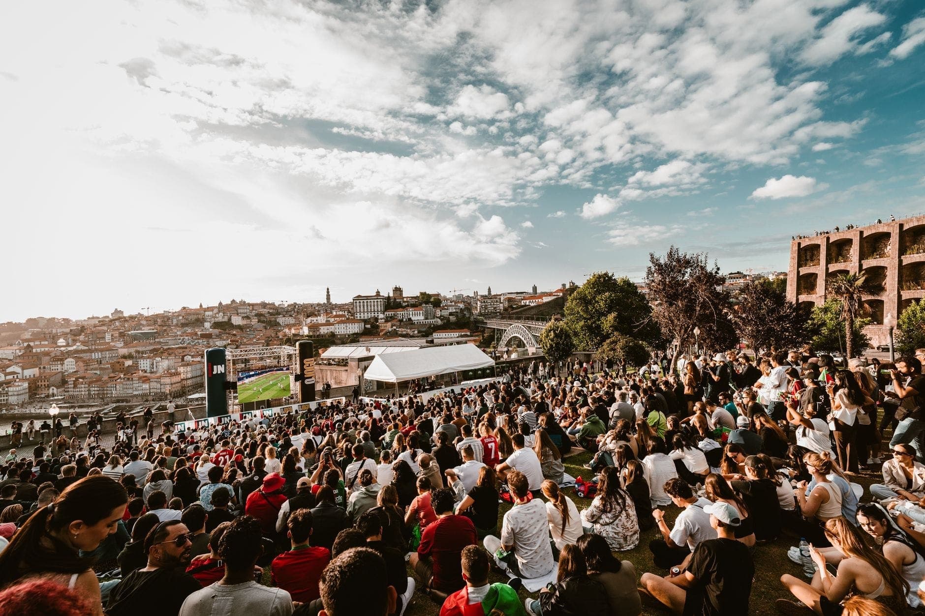 Gran multitud de personas sentadas en una ladera cubierta de hierba con la ciudad de Porto y el río Duero al fondo bajo un cielo parcialmente nublado.
