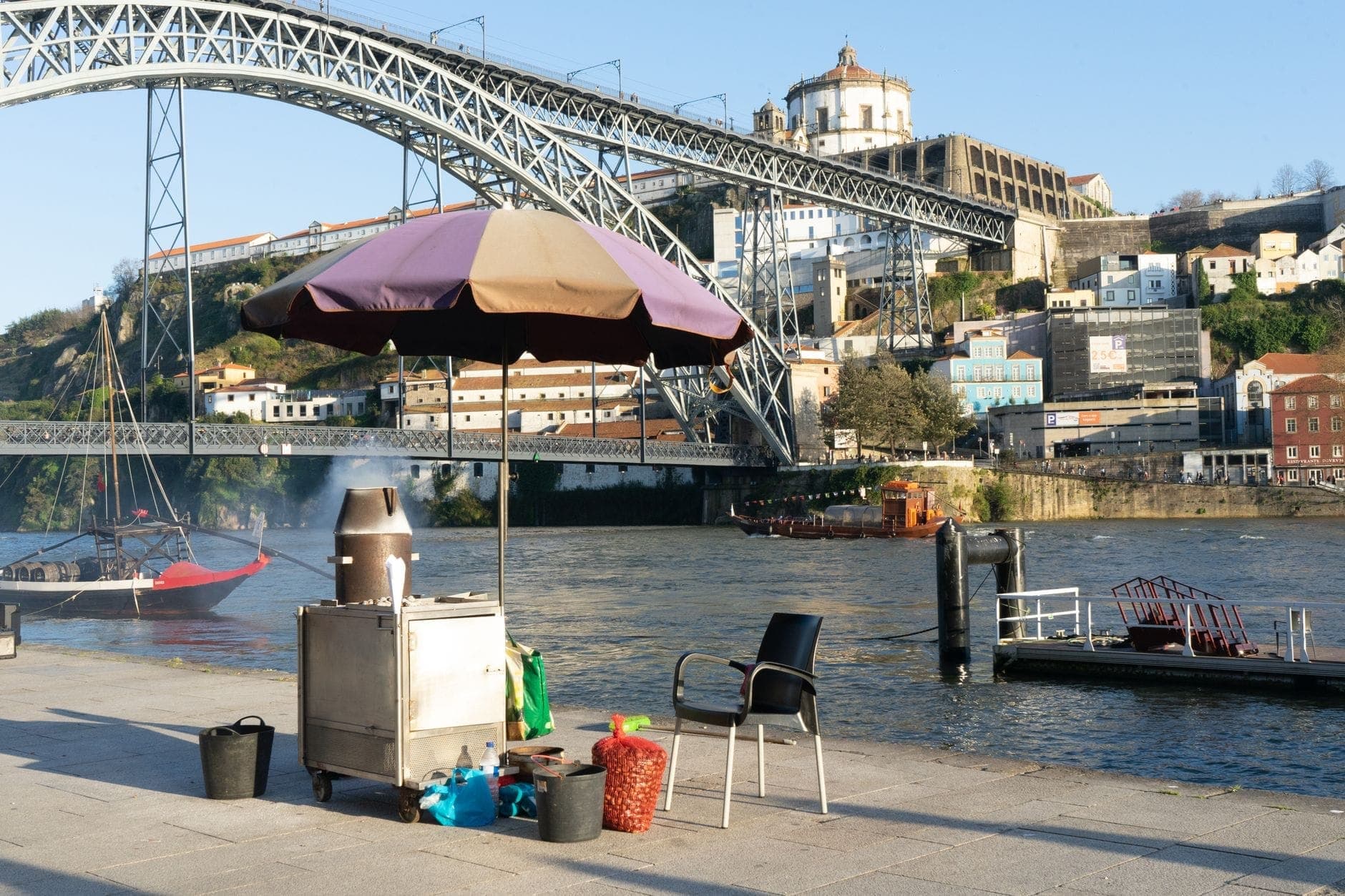 Escena junto al río en Oporto con un carrito de comida callejera y parrilla junto al río Duero, con el icónico puente y la histórica ciudad al fondo.