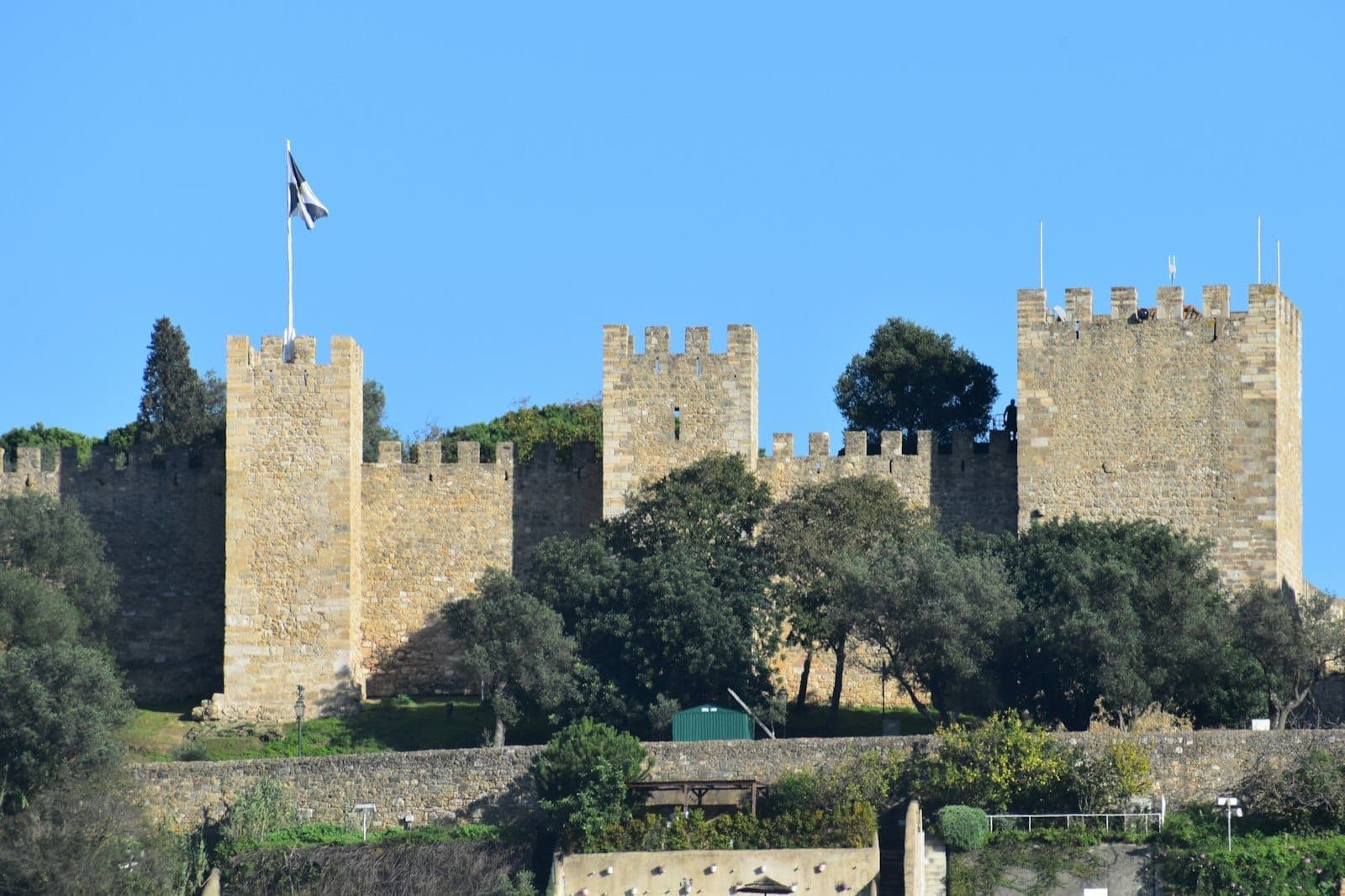Torres de pedra e muralhas fortificadas do Castelo de São Jorge erguem-se acima de árvores verdes sob um céu azul claro em Lisboa, Portugal.
