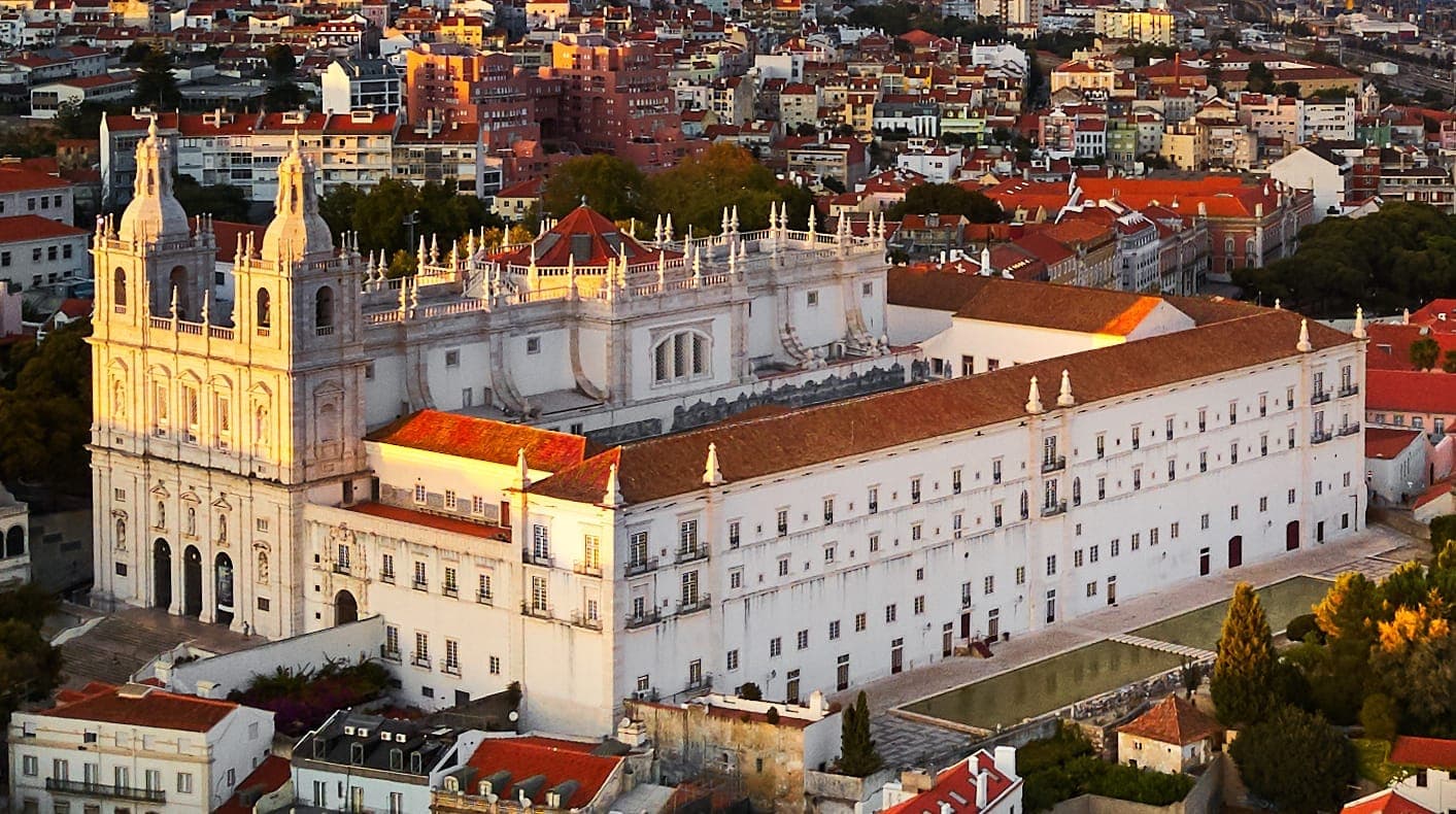 Vista aérea da Igreja de São Vicente de Fora com sua fachada barroca branca, telhado de telhas vermelhas e a paisagem urbana de Lisboa ao pôr do sol.