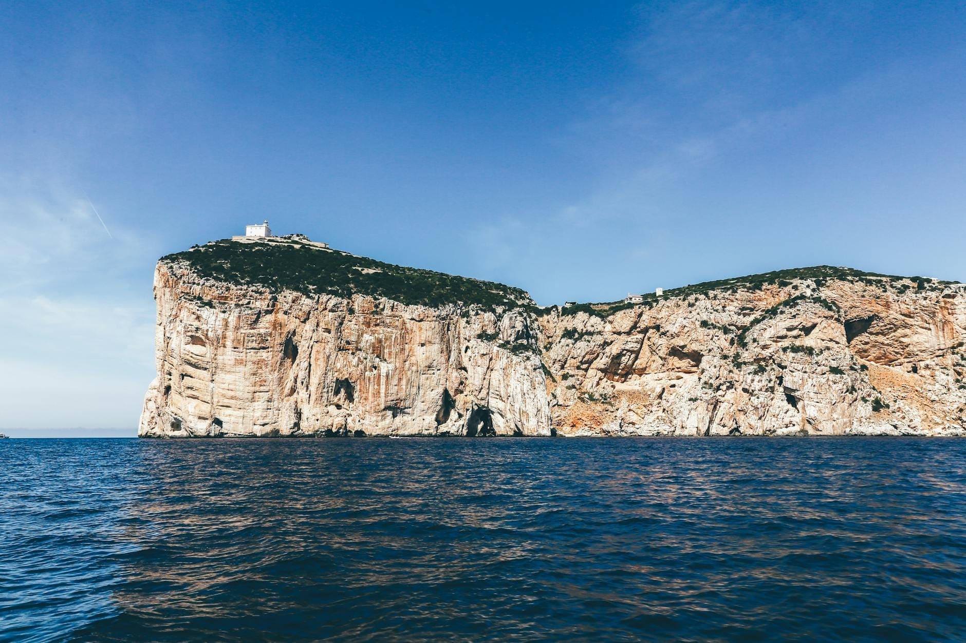 Klippen von Capo Caccia mit einem Wachturm, die sich über das tiefblaue Meer an der Nordwestküste Sardiniens unter einem klaren blauen Himmel erheben.
