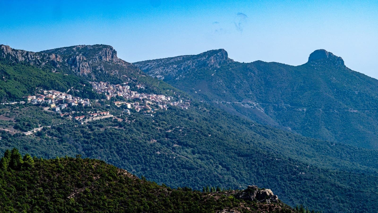 Distant view of a Sardinian mountain village nestled in green hills with rugged peaks in the background under a clear blue sky.