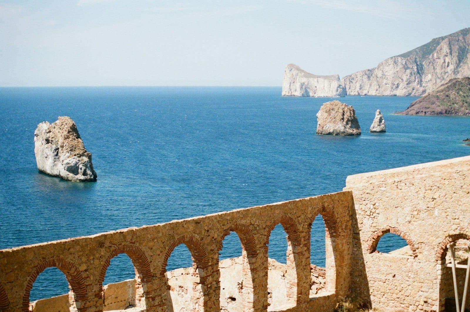 Dramatic limestone sea stacks and cliffs rising from the blue Mediterranean, viewed behind stone arches of an old mining structure on the Sardinian coast.