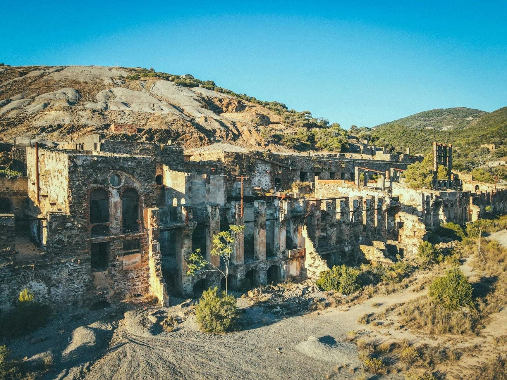 Ruins of an old mining complex in arid hills, with dramatic stone buildings and natural landscape under a blue sky.