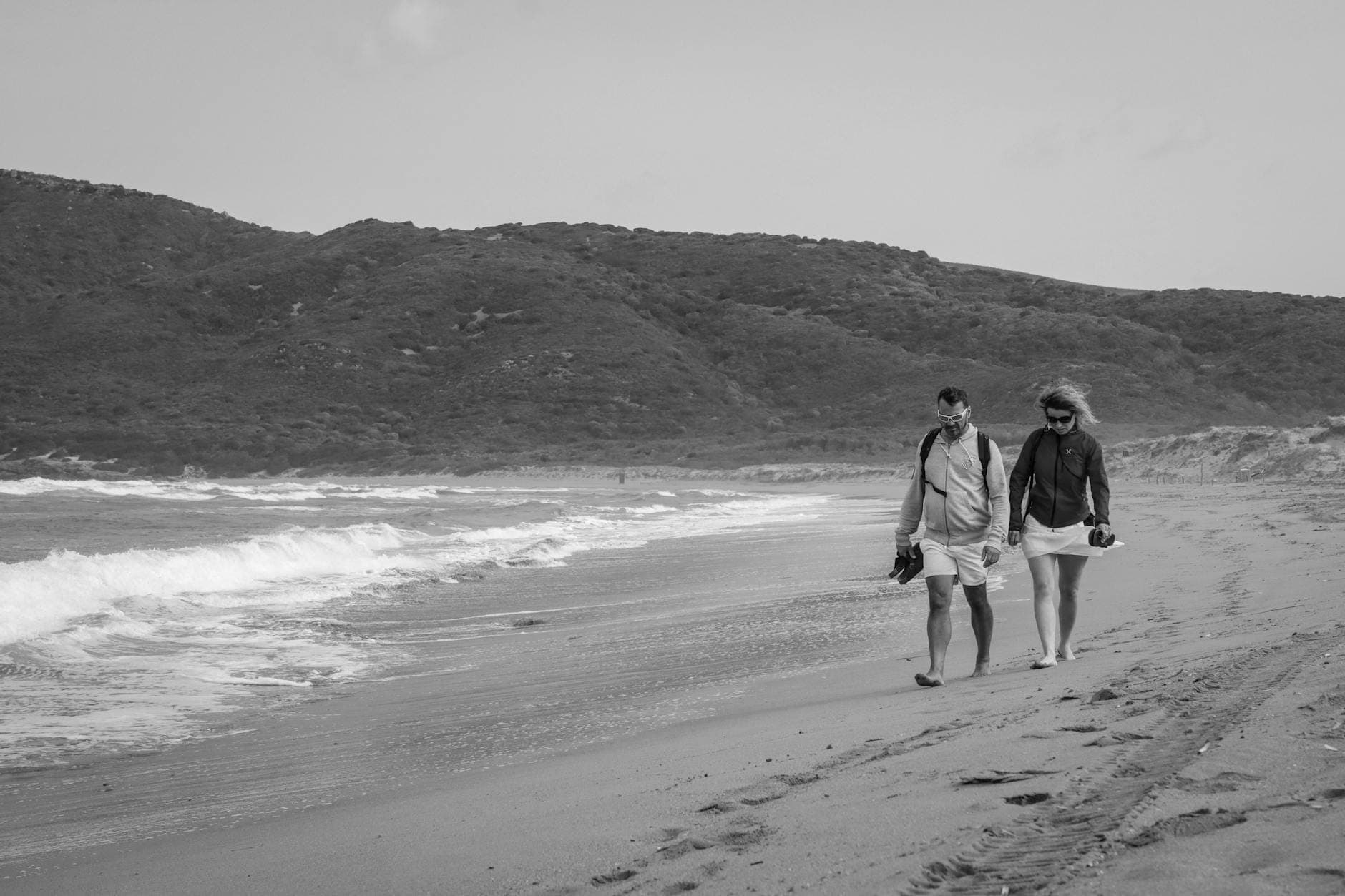 Una pareja camina de la mano por una playa de arena con olas suaves y colinas verdes al fondo.