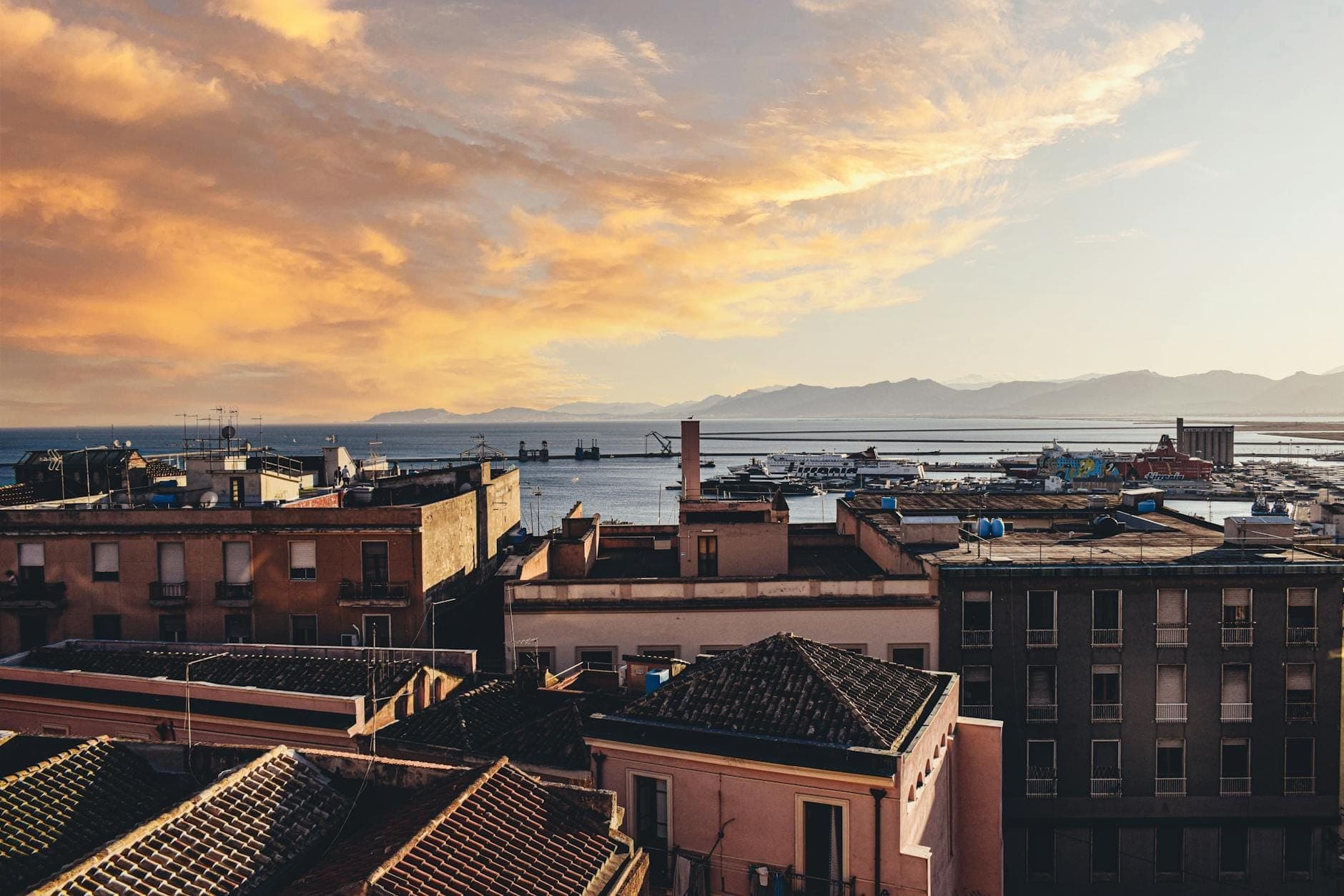 Vista desde la azotea de una ciudad costera de Cerdeña al atardecer, con el puerto, el paseo marítimo y edificios en colores pastel bajo un cielo naranja.