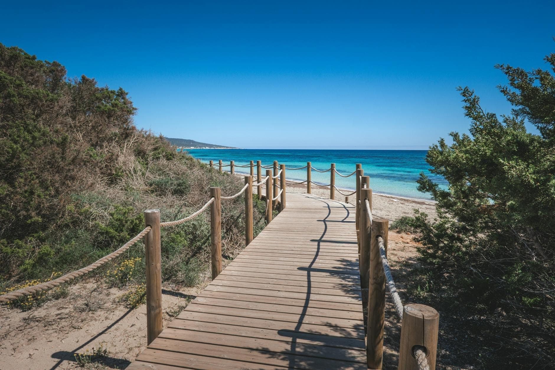 Pasarela de madera que lleva hacia una playa de arena y el mar turquesa bajo un cielo azul despejado, rodeada de arbustos mediterráneos verdes.