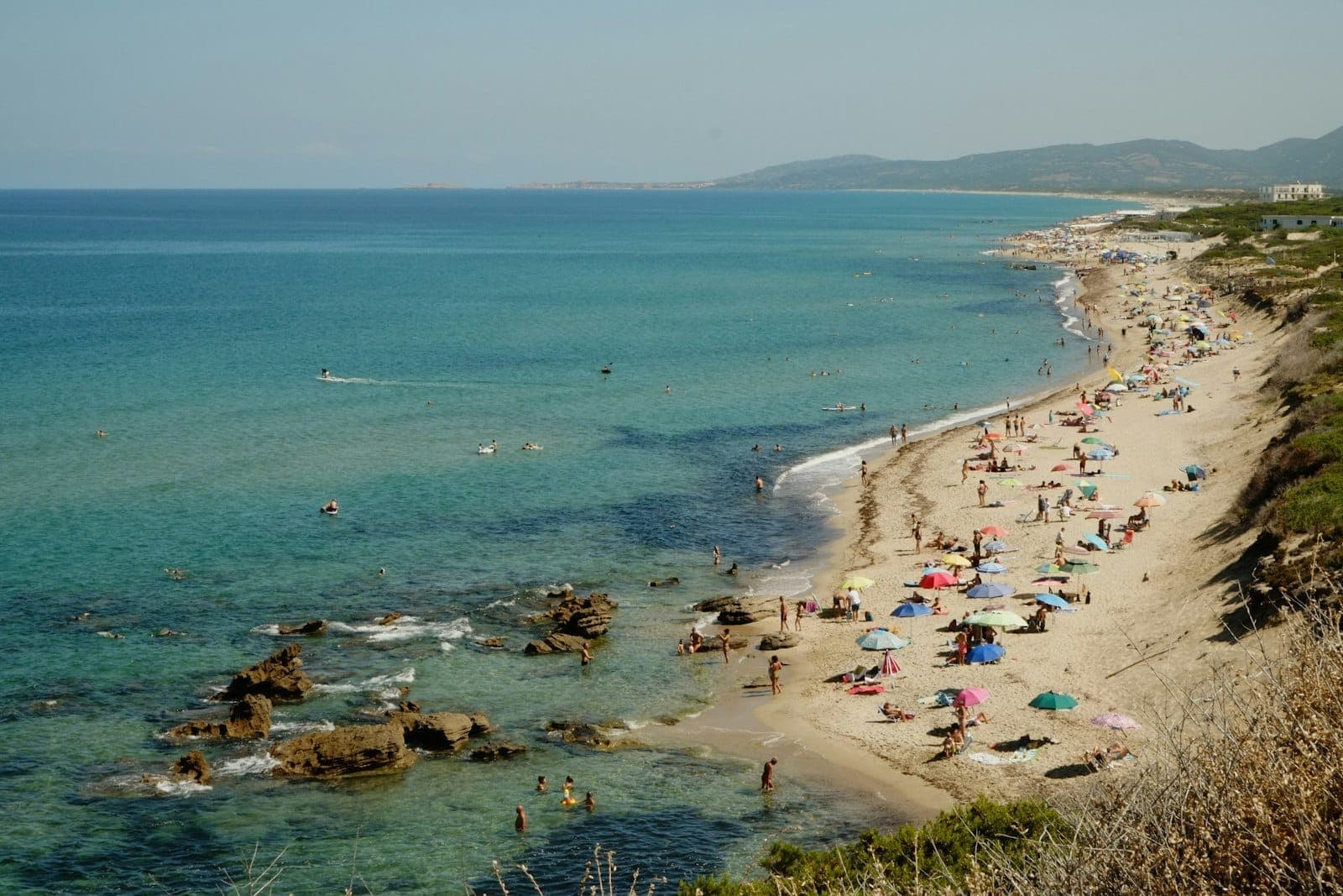 Larga playa de arena en Cerdeña con mar turquesa, bañistas, sombrillas de colores y cielos despejados de verano.
