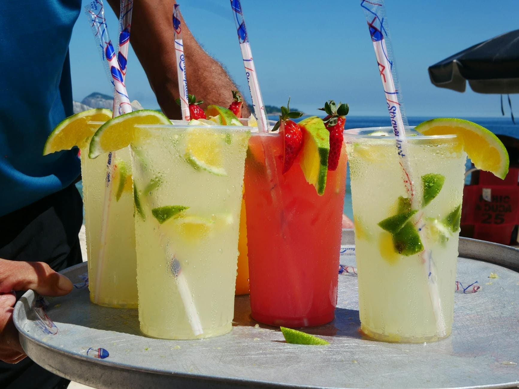 Primer plano de vasos de plástico llenos de coloridas bebidas sin alcohol decoradas con limón y frutas en una bandeja en la playa.