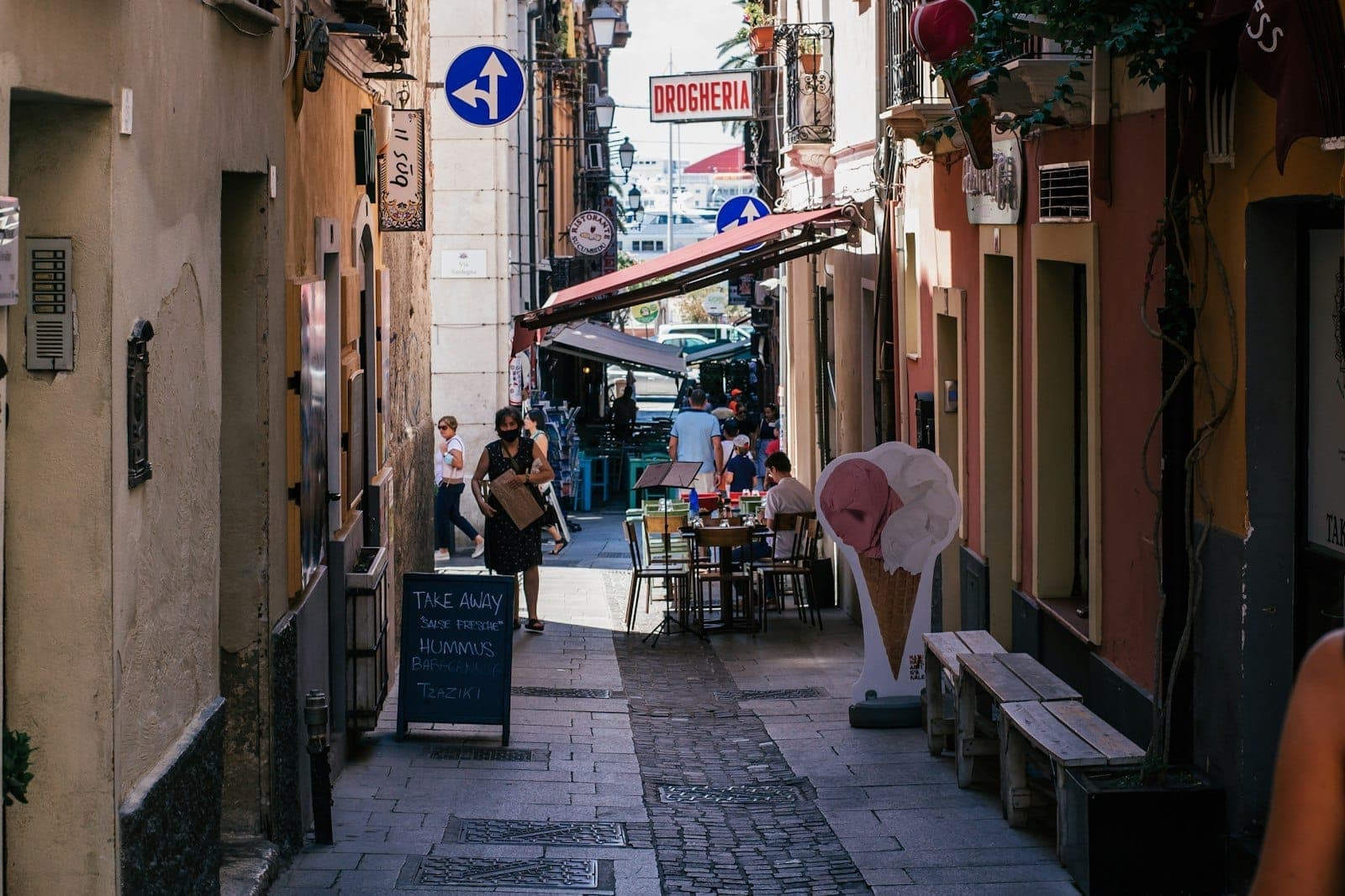Calle estrecha y animada de Cerdeña con pequeñas mesas, personas sentadas al aire libre, letreros de restaurantes y típicos edificios mediterráneos.