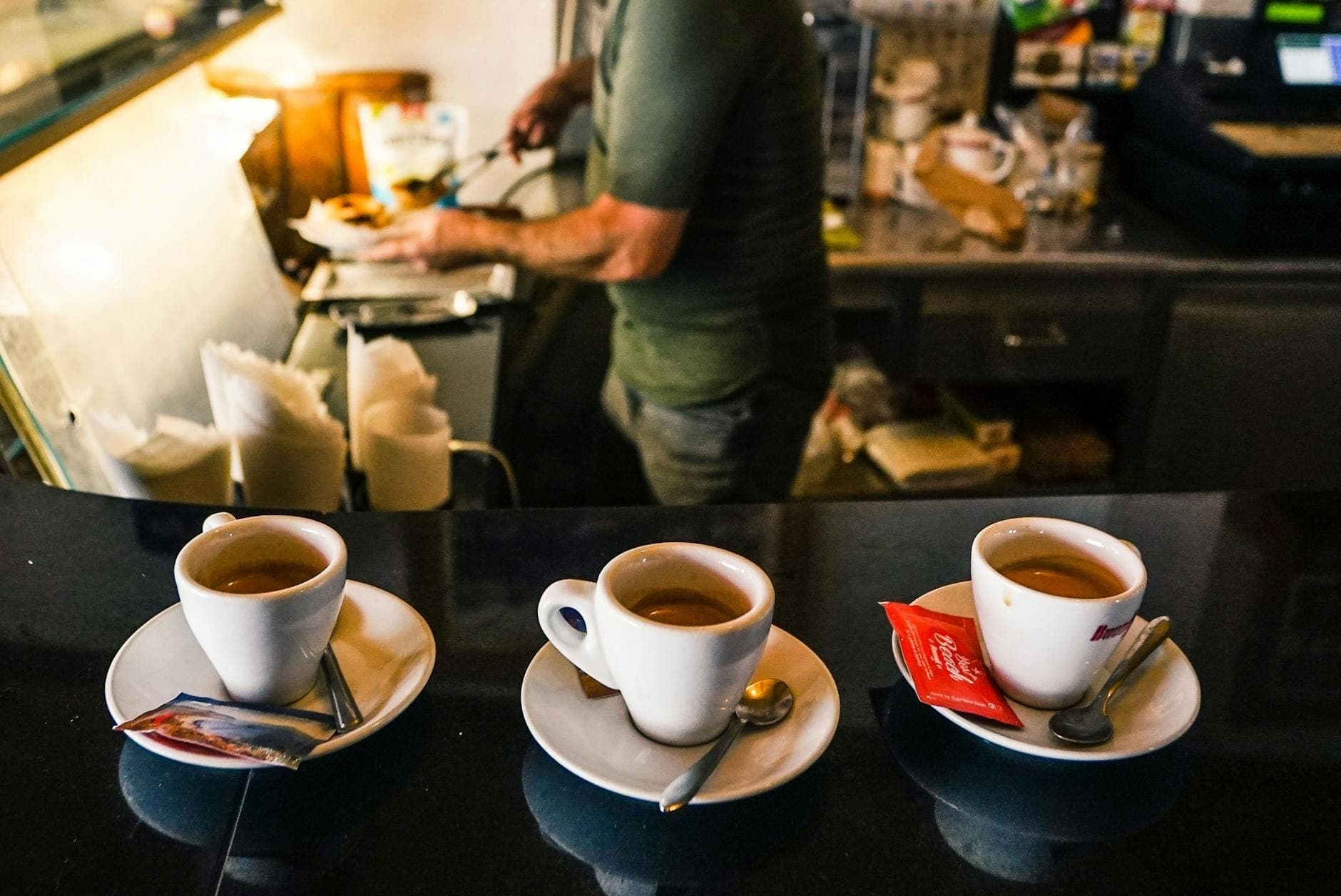 Tres tazas de espresso en la barra de un café con un barista preparando bebidas al fondo, evocando la cultura del café italiano.