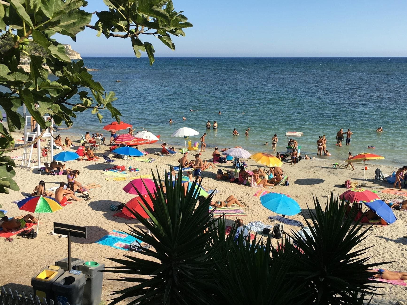 Überfüllte Strandszene auf Sardinien mit bunten Sonnenschirmen, Sonnenbadenden und Schwimmern, die Hochsaison und Planungsbedarf heraufbeschwören.