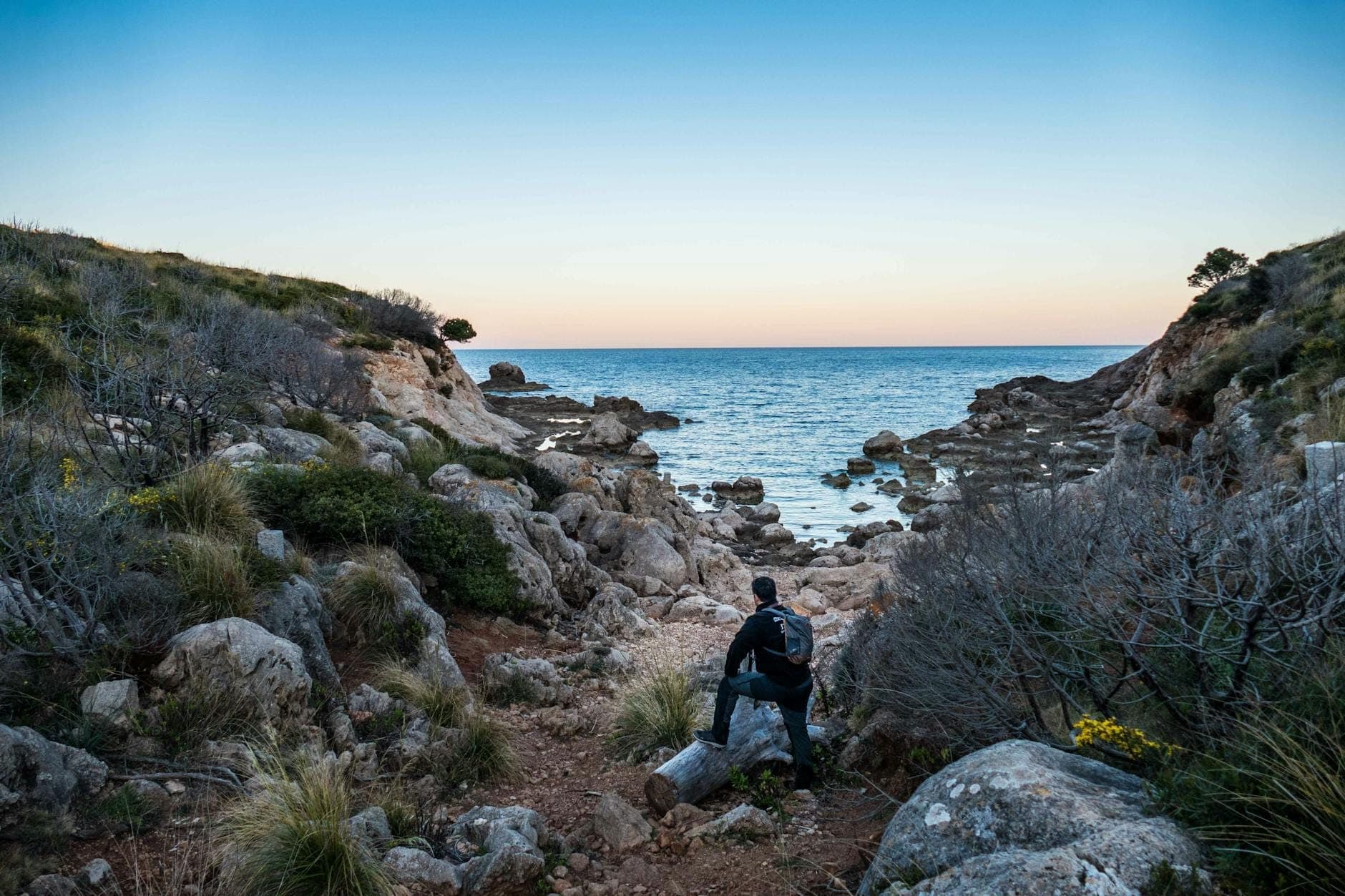 Ein Wanderer mit Rucksack sitzt auf einem Baumstamm mit Blick auf eine felsige Bucht und das blaue Mittelmeer bei Sonnenuntergang auf Sardinien.