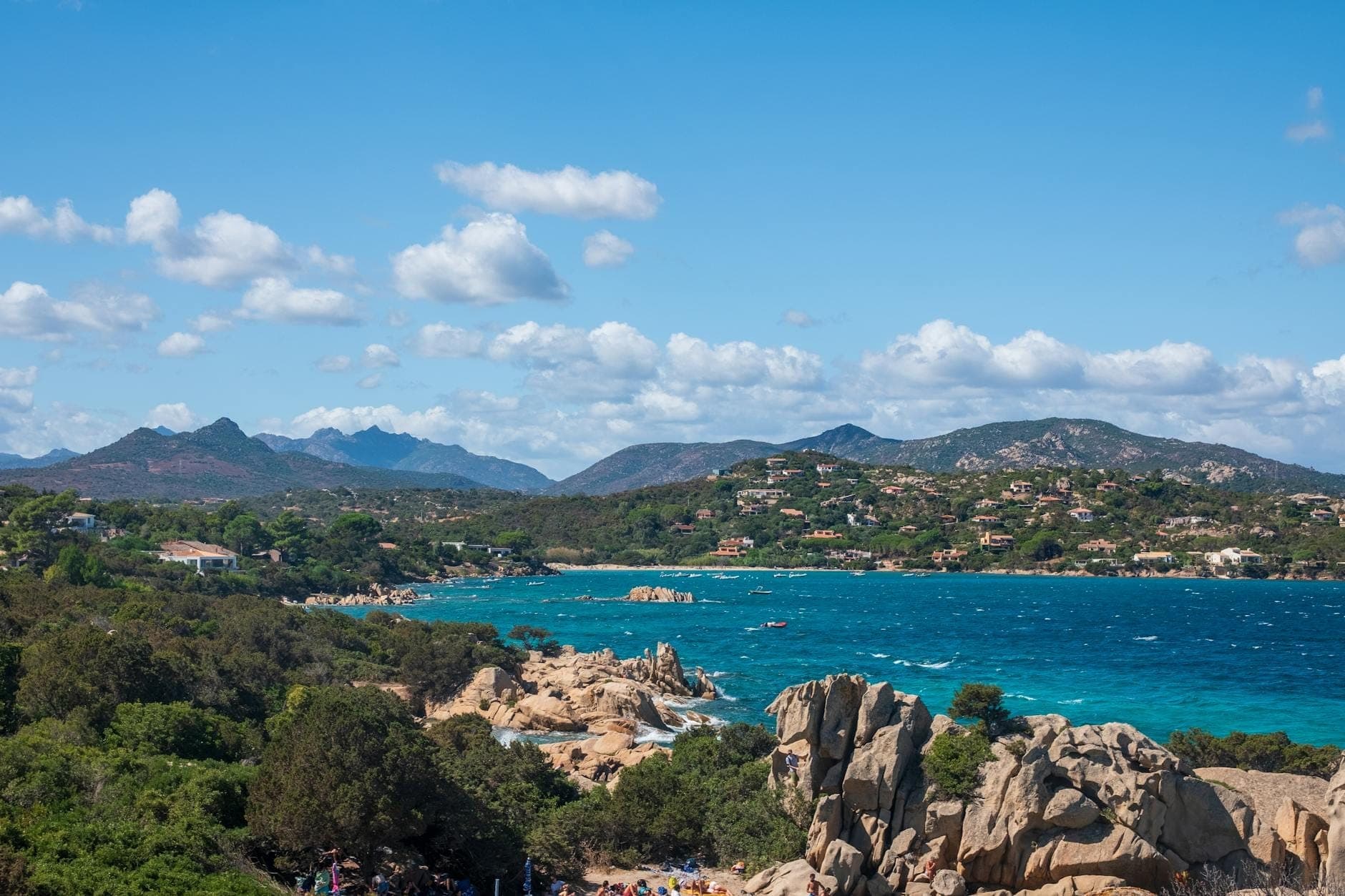 Panoramablick auf die sardische Küste mit türkisblauem Meer, felsigem Ufer, üppigem Grün, fernen Hügeln und vereinzelten Wolken an einem strahlend sonnigen Tag.