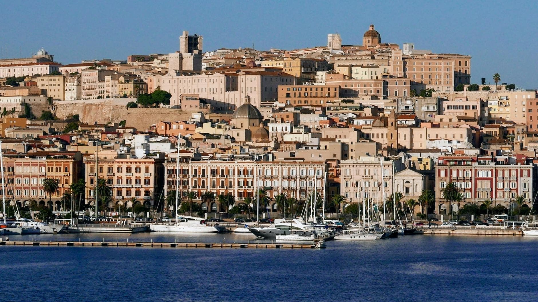 Panoramablick auf die Uferpromenade von Cagliari, der südlichen Stadt Sardiniens, mit sonnenbeschienenen historischen Gebäuden, die sich auf einem Hügel über einem Yachthafen mit vertäuten Jachten stapeln.