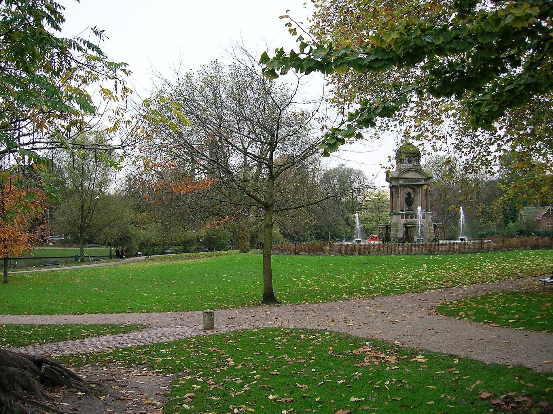 Weitwinkelansicht des Sarphatipark mit üppigen grünen Rasenflächen, Gehwegen, verstreuten Herbstblättern und dem historischen Steindenkmal mit Brunnen im Hintergrund.