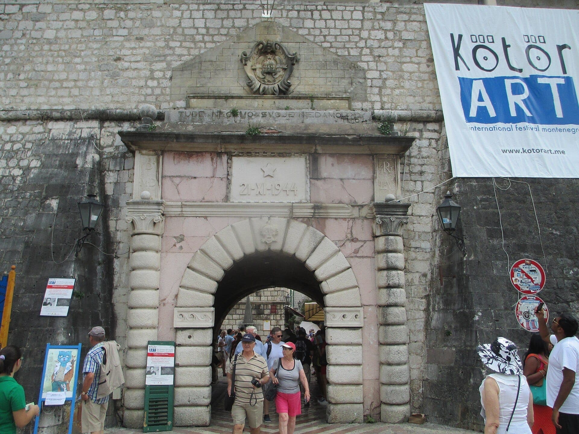 Des visiteurs franchissent l'historique arche en pierre de la Porte de Mer de Kotor, ornée de reliefs et encadrée par les anciens remparts de la vieille ville.