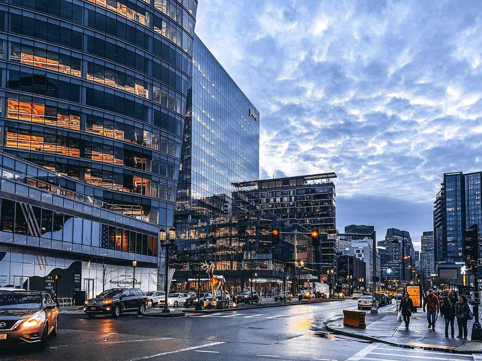 Des immeubles modernes en verre longent une rue animée du Seaport District de Boston au crépuscule, avec des voitures et des piétons se déplaçant sous un ciel nuageux spectaculaire.