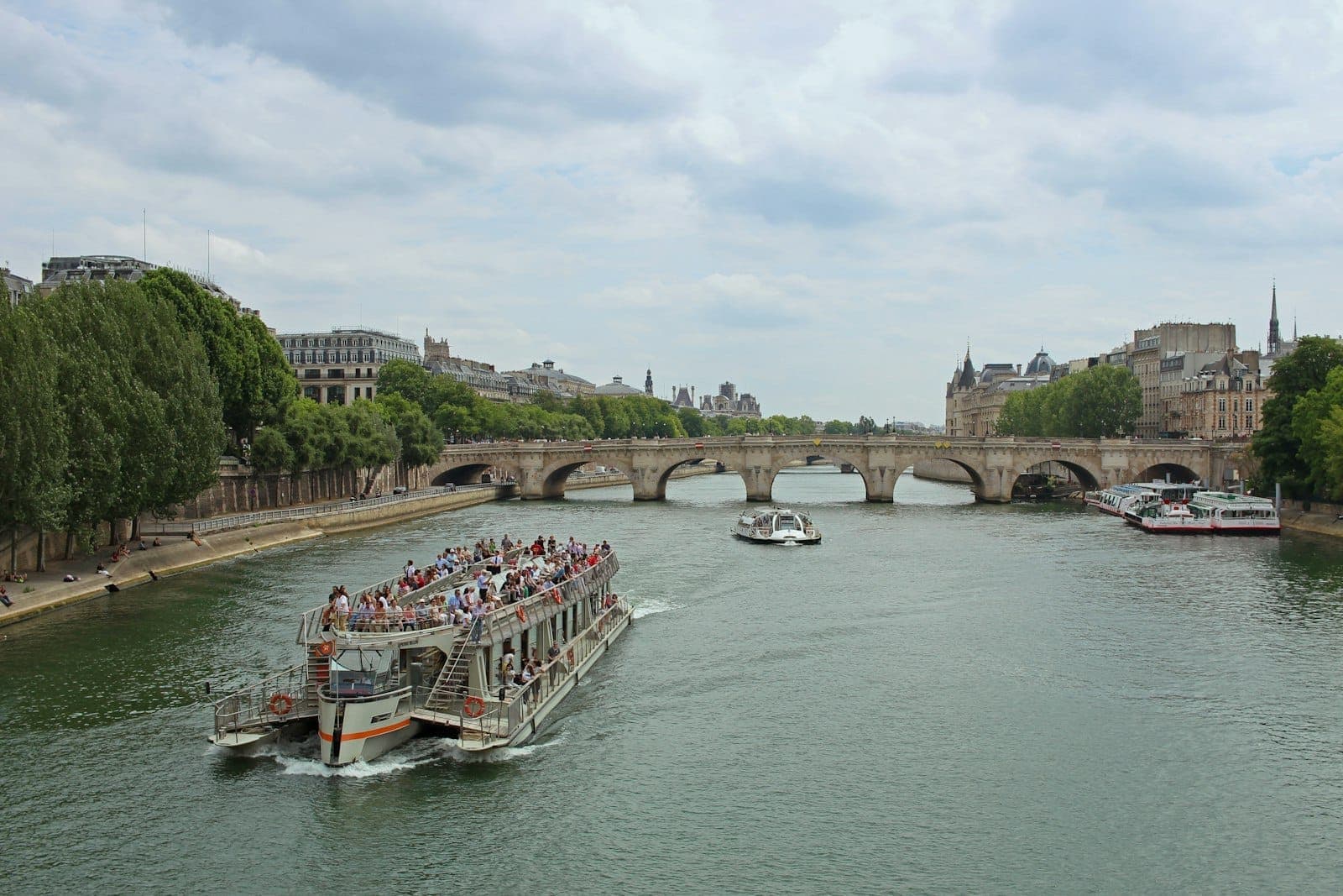 A river cruise boat filled with tourists glides along the Seine in Paris, passing under historic bridges and tree-lined banks on a bright day.