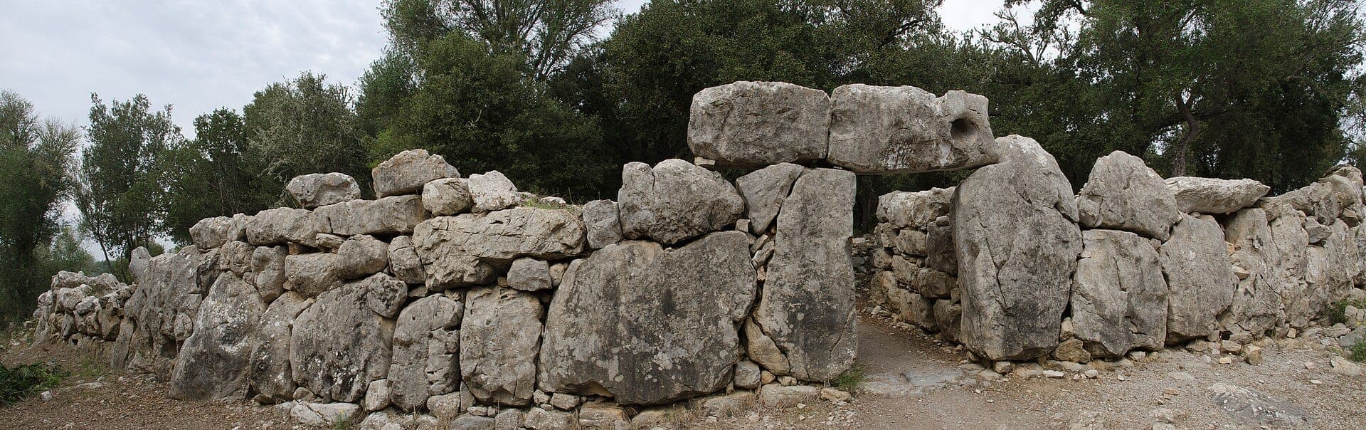 Panoramablick auf die antiken zyklopischen Steinmauern der archäologischen Stätte Ses Païsses, umgeben von grünen Steineichen unter bewölktem Himmel.