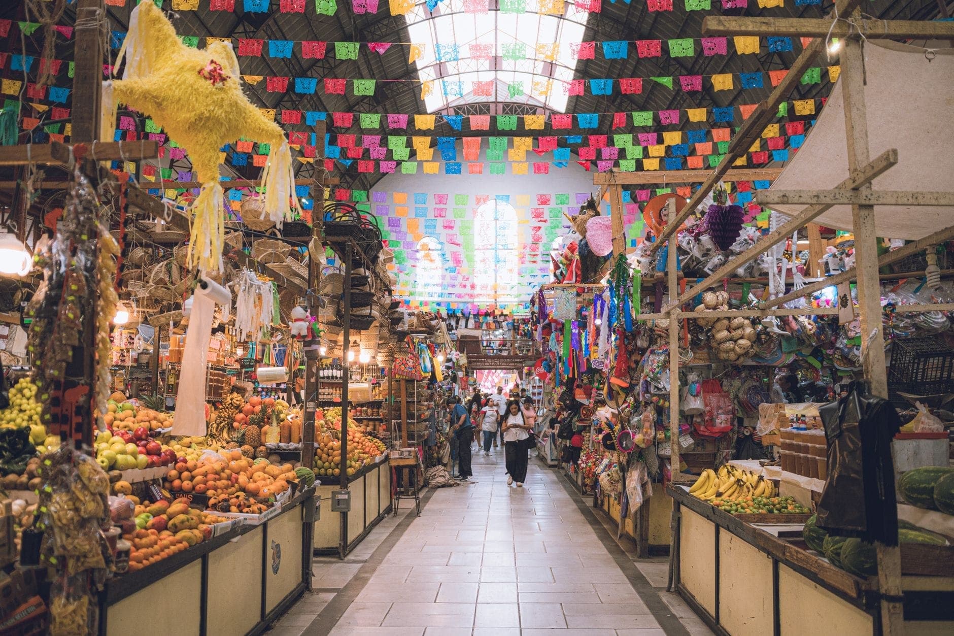 ภายในของ Mercado Libertad ที่มีสีสันสดใส พร้อมแผงขายผลไม้และสินค้า การตกแต่งด้วย papel picado และแสงธรรมชาติที่ส่องผ่านหลังคา
