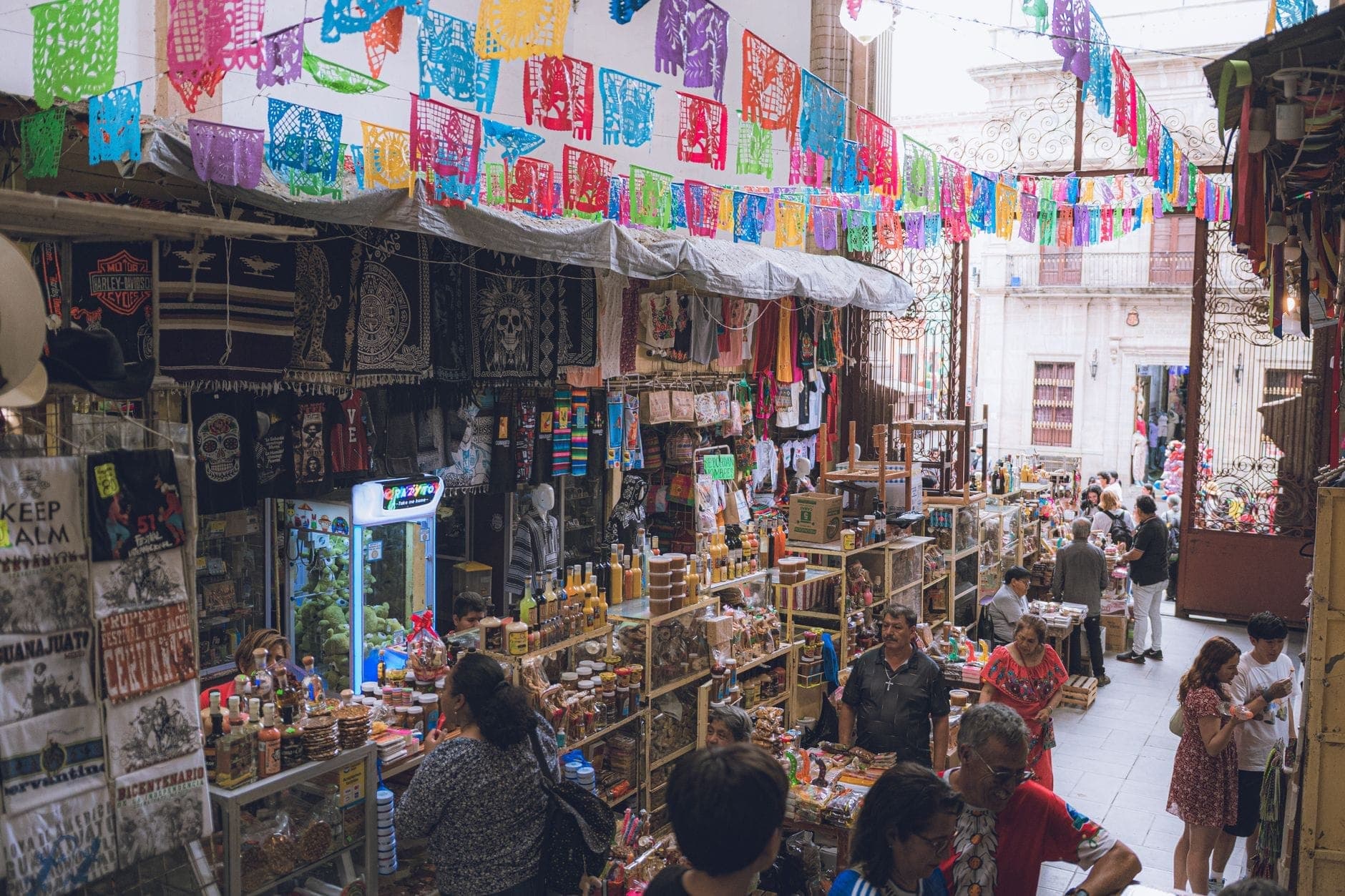 ตลาดในร่มที่กวาดาลาฮารา เต็มไปด้วยสินค้างานฝีมือสีสันสดใส แขวนด้วยกระดาษ papel picado และผู้คนกำลังเลือกซื้อจากแผงต่าง ๆ