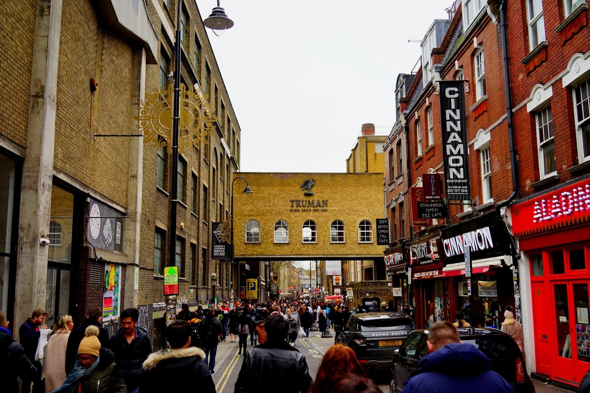 Multitudes caminan por una concurrida calle en Shoreditch, East End, enmarcadas por edificios de ladrillo y el histórico letrero de Truman Brewery en un día nublado.
