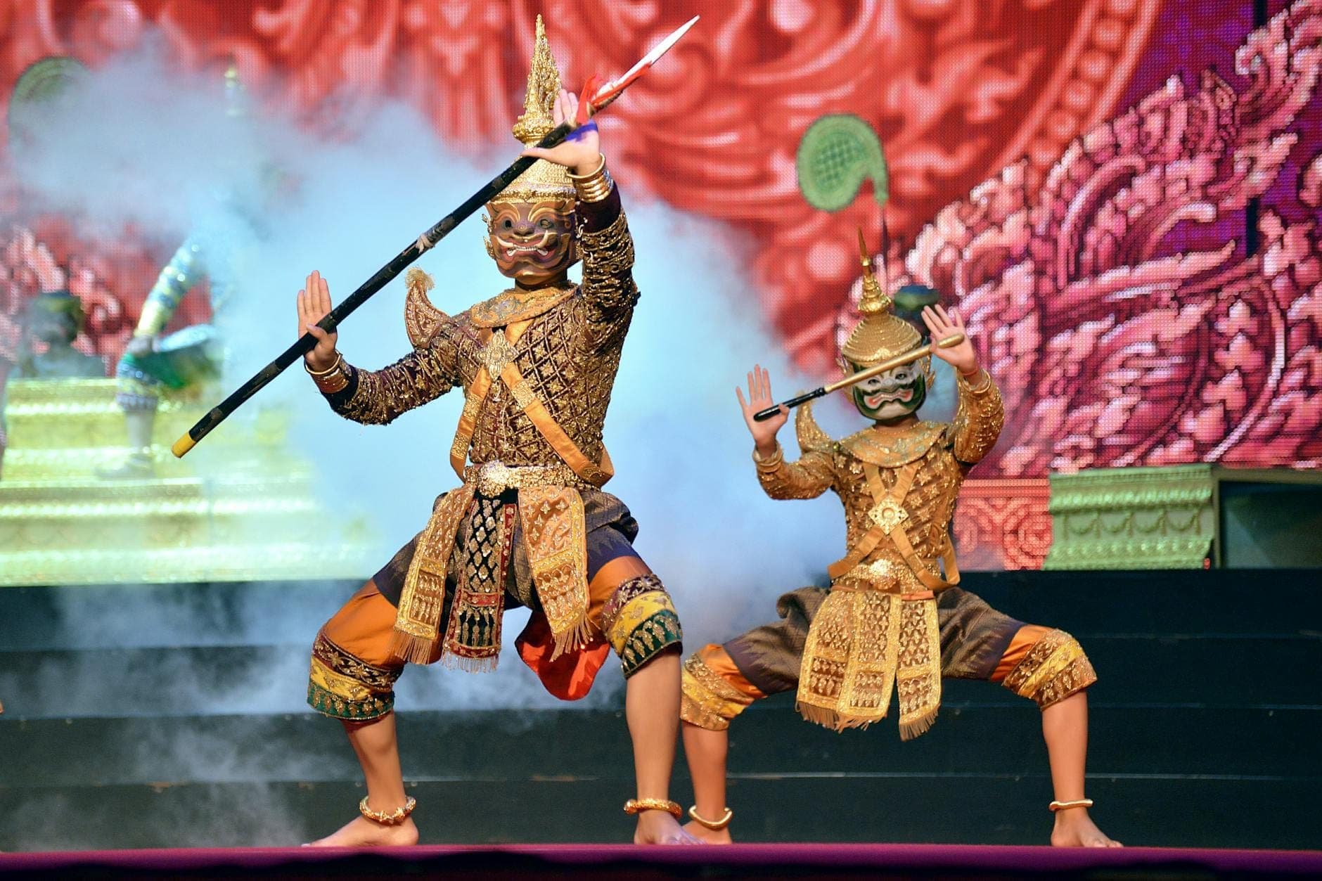 Two performers in traditional Thai costumes and masks pose dramatically on stage at Siam Niramit Phuket, with vibrant patterns and theatrical lighting.