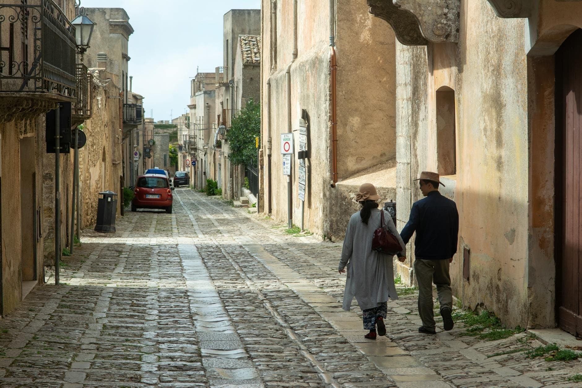 Una coppia mano nella mano passeggia lungo una tranquilla strada acciottolata e assolata, fiancheggiata da affascinanti edifici storici in Sicilia.