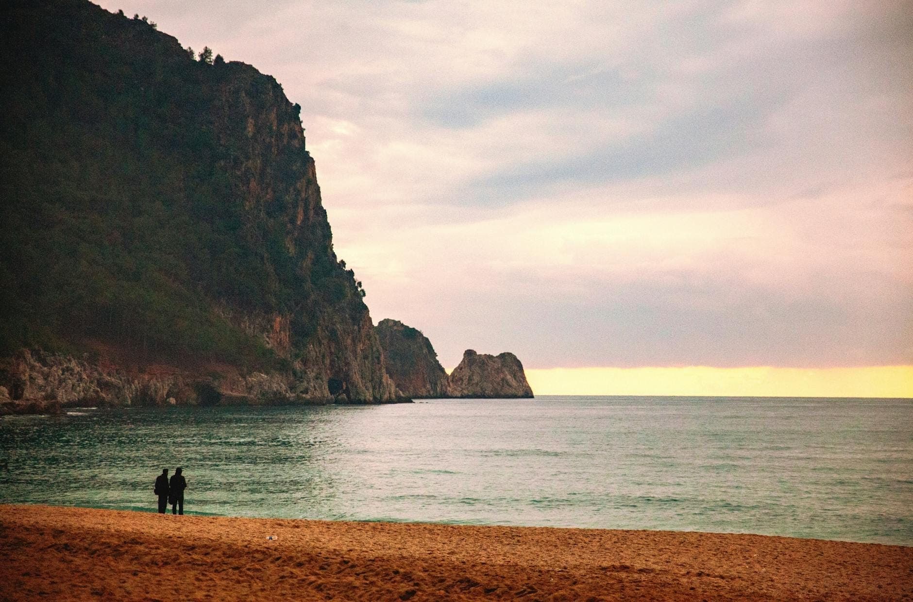 Una coppia si trova insieme su una spiaggia sabbiosa al tramonto, con lo sguardo verso il mare e spettacolari scogliere rocciose sullo sfondo.
