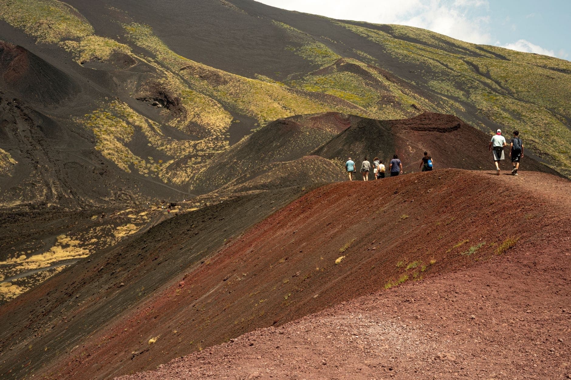 Gruppo di persone che fanno trekking lungo una cresta vulcanica rossa sull'Etna con un drammatico paesaggio vulcanico sullo sfondo.