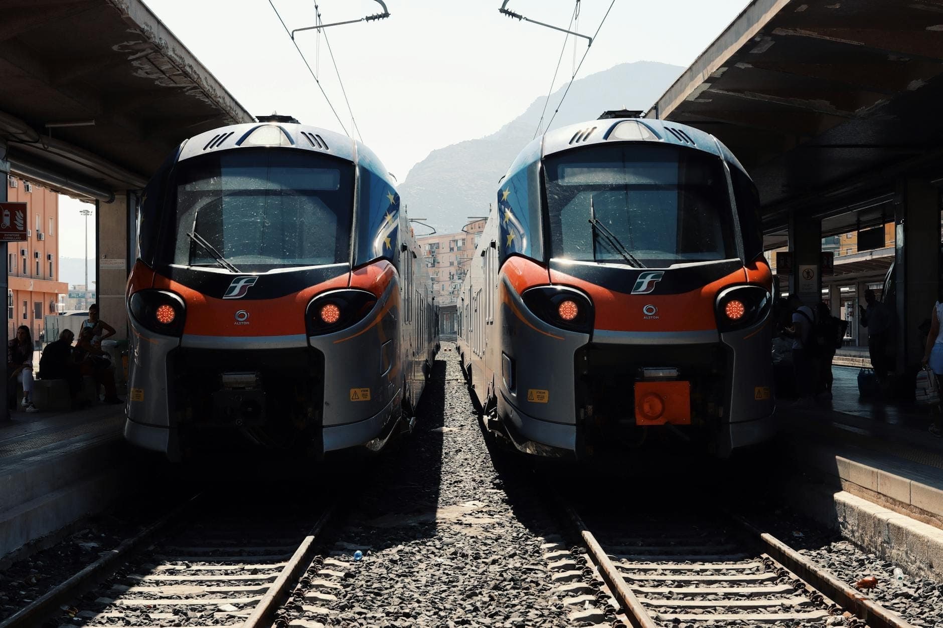 Dua kereta modern di peron stasiun di Sisilia, dengan pegunungan terlihat di latar belakang dan para pelancong menunggu di dekatnya.