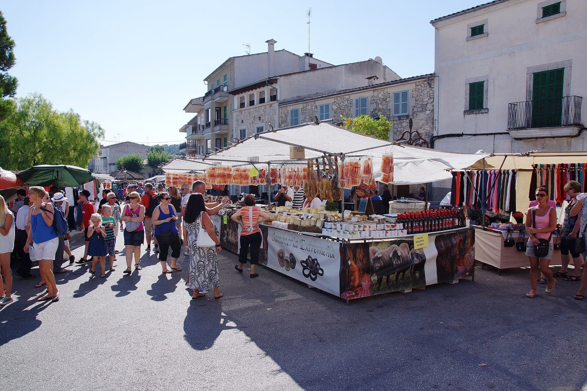 Menschenmengen durchstöbern Freiluftmarktstände mit lokalen Erzeugnissen, Fleisch und Kunsthandwerk auf dem Sineu Mittwochsmarkt, mit traditionellen Steingebäuden im Hintergrund.