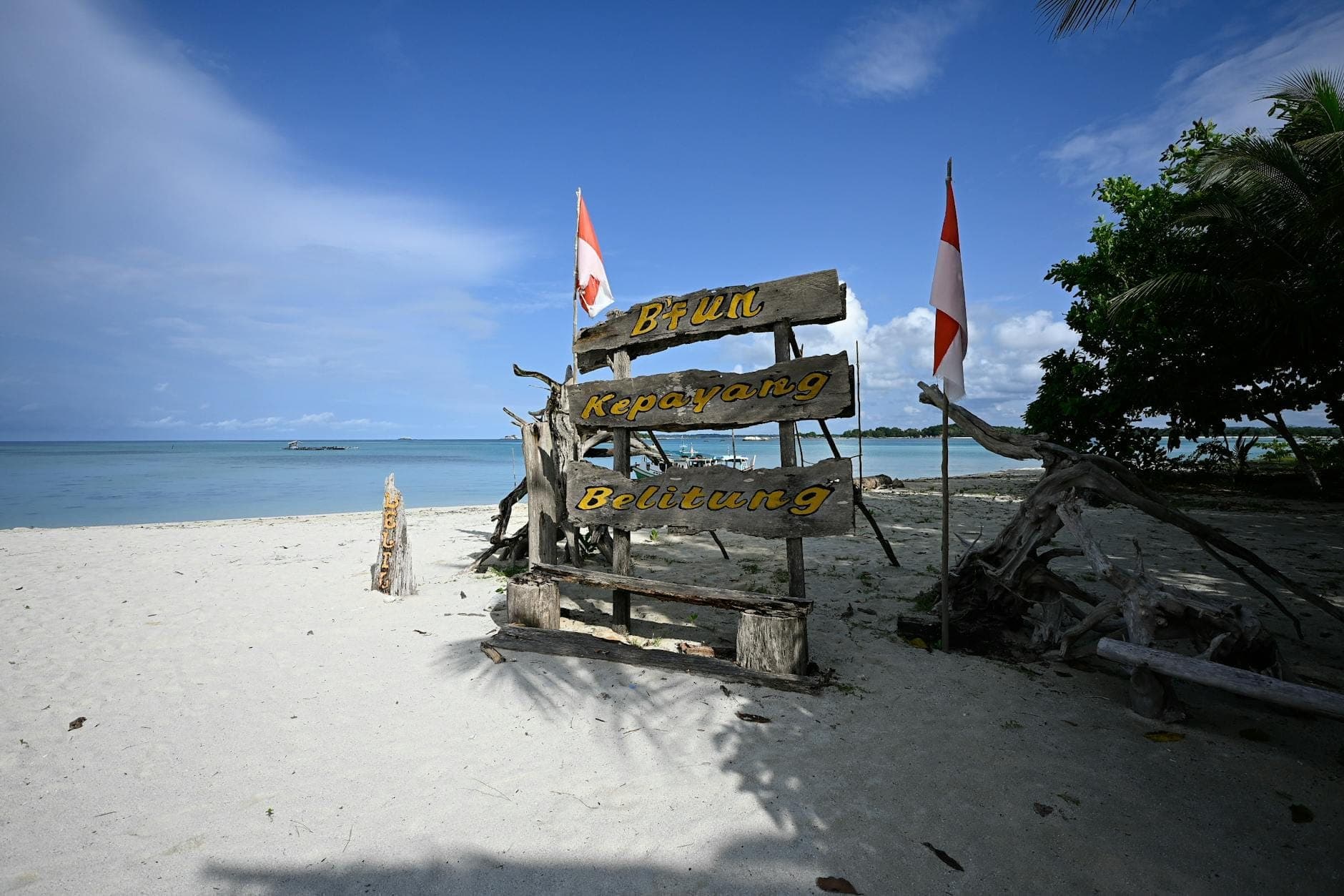 Sandstrand mit Holzschild mit der Aufschrift Bintan, Kepatang, Belitung und zwei indonesischen Flaggen am Meer unter blauem Himmel