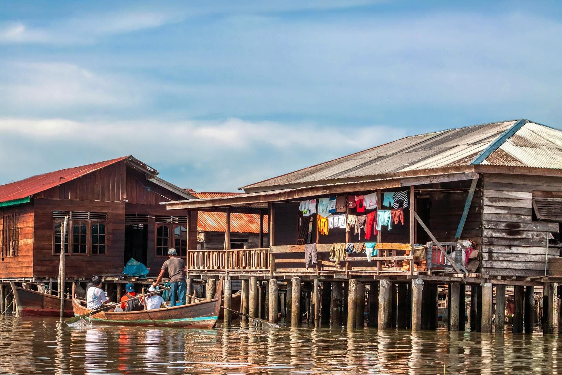 Hölzerne Pfahlbauten über dem Wasser mit hängender Wäsche, ein kleines Boot mit Menschen davor, blauer Himmel im Hintergrund.
