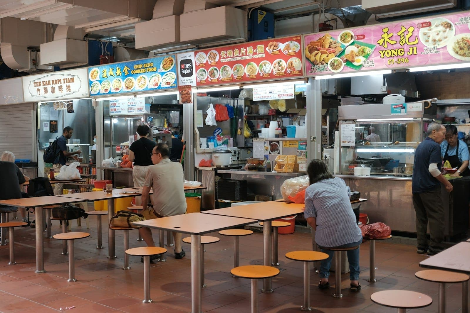 Innenraum eines Hawker Centre in Singapur mit Essensständen, Speisekarten oben und Menschen, die an Gemeinschaftstischen speisen.