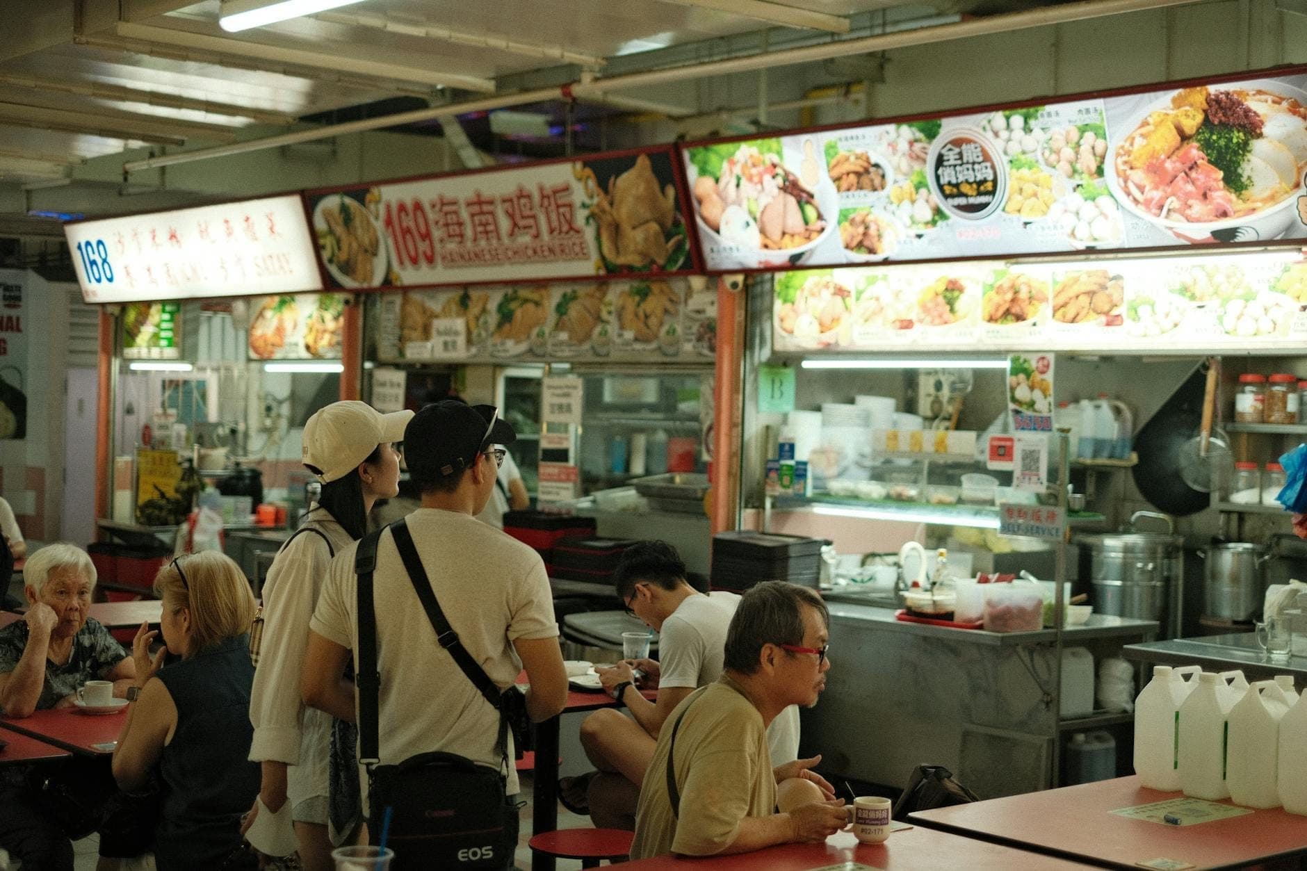 Menschen stehen Schlange und speisen in einem hell erleuchteten Hawker Centre in Singapur mit Essensständen und Speisekarten an der Decke.