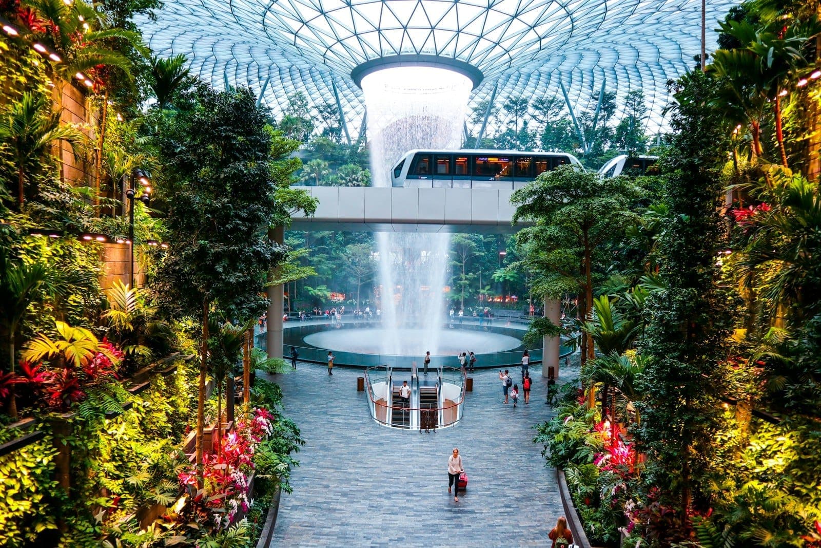 มุมมองกว้างของน้ำตกในร่มของ Jewel Changi Airport ที่둘러มด้วยพืชพรรณเขียวชอุ่ม พื้นที่เดินเล่นโล่ง และรางรถไฟ Skytrain เชื่อมต่อที่อยู่ด้านบน