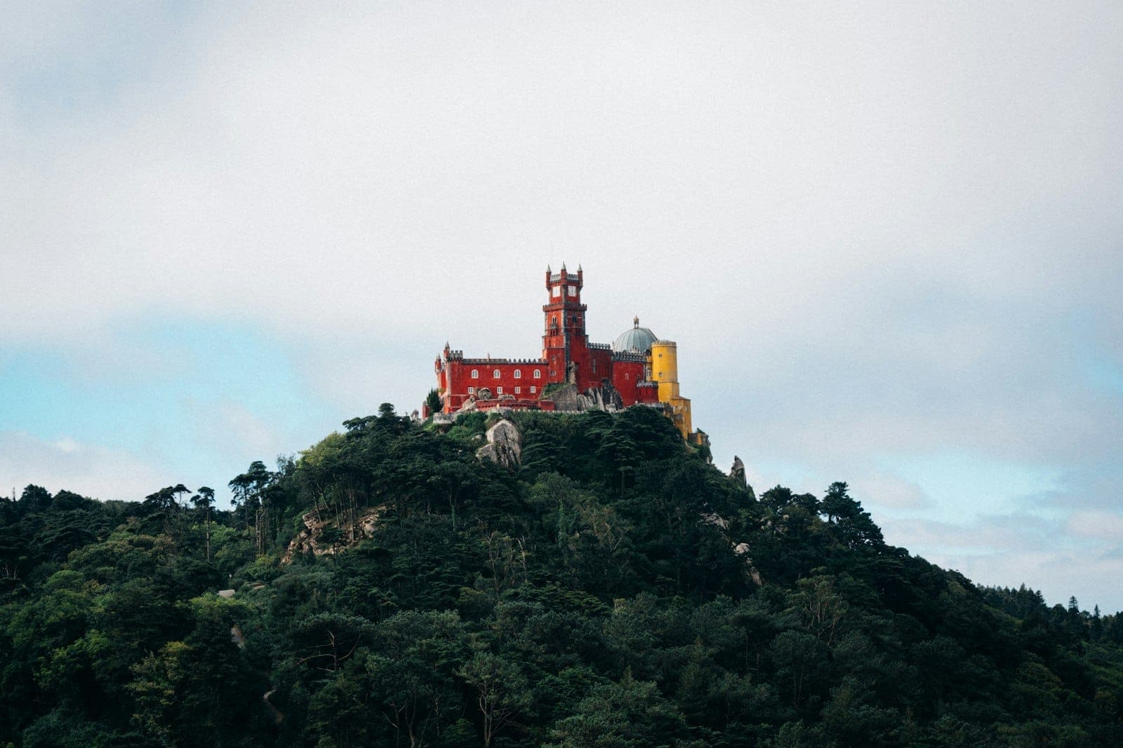 O colorido Palácio da Pena no alto de uma exuberante colina arborizada em Sintra, sob um dramático céu nublado.