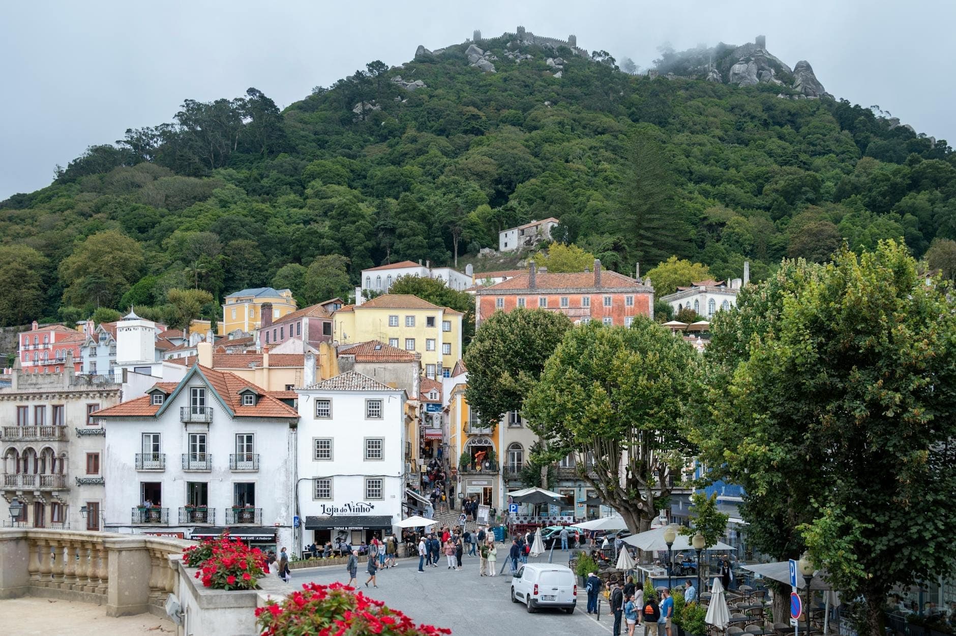 Vista do centro de Sintra com edifícios coloridos, pedestres, carros e o Castelo dos Mouros na colina arborizada acima.