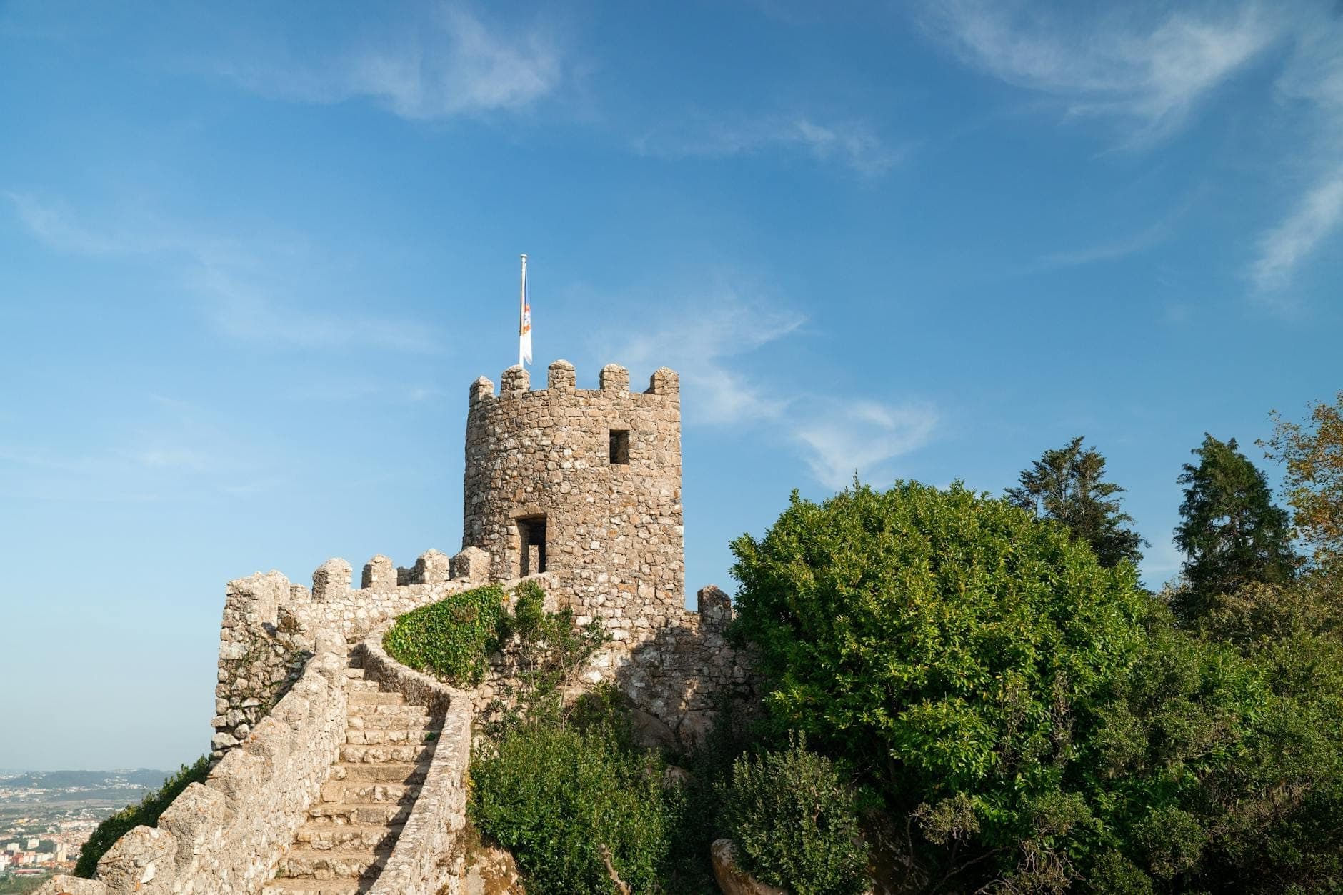 Degraus de pedra serpenteando até as ameias do Castelo dos Mouros em Sintra com céu azul e vegetação.