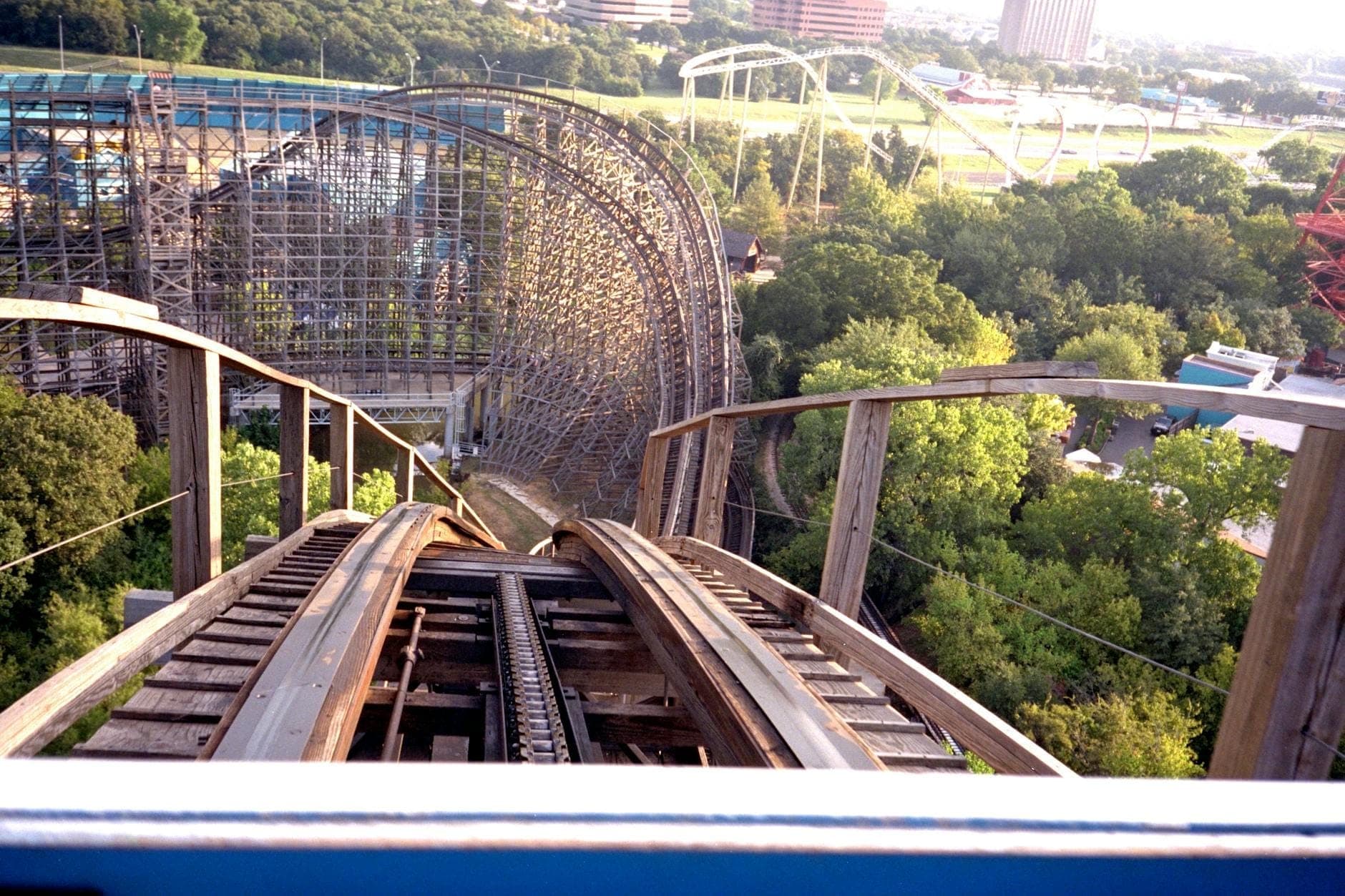 Una emocionante toma en primera persona desde lo alto de una montaña rusa de madera, rodeada de árboles verdes y estructuras del parque al fondo.