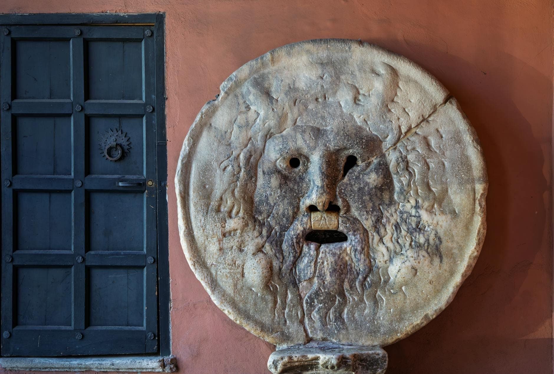 Vista ravvicinata del famoso volto in pietra della Bocca della Verità a Roma accanto a una porta scura.